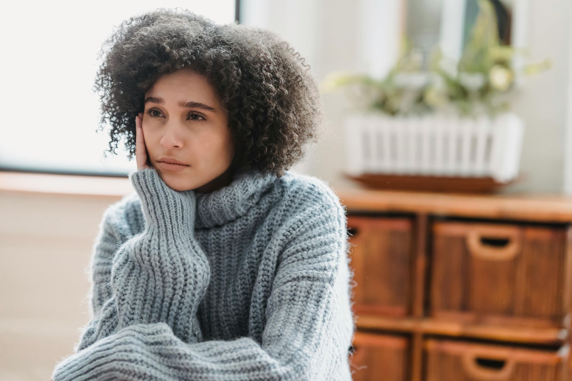 Woman with curly hair, resting her head on her hand, appears sad in a room.