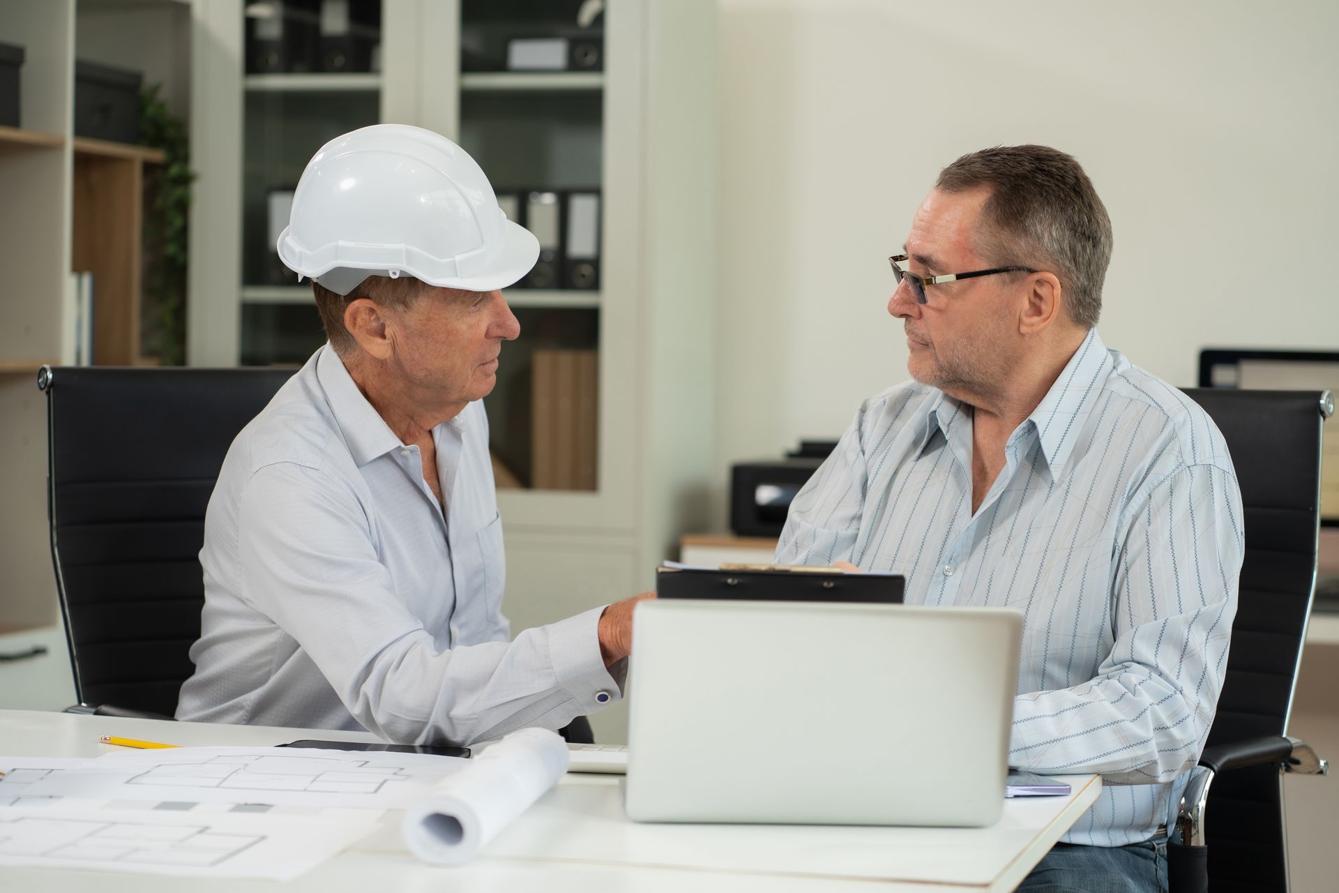Two men in office, one wearing a hard hat, reviewing papers and laptop.