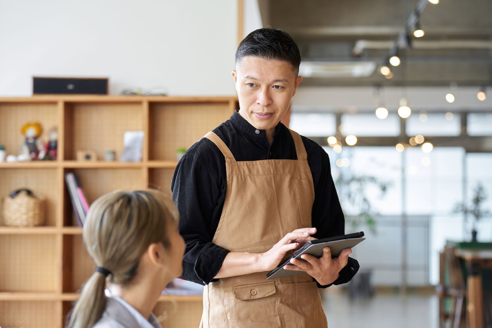 Man in apron uses tablet, speaking to a person at a table in a cafe.