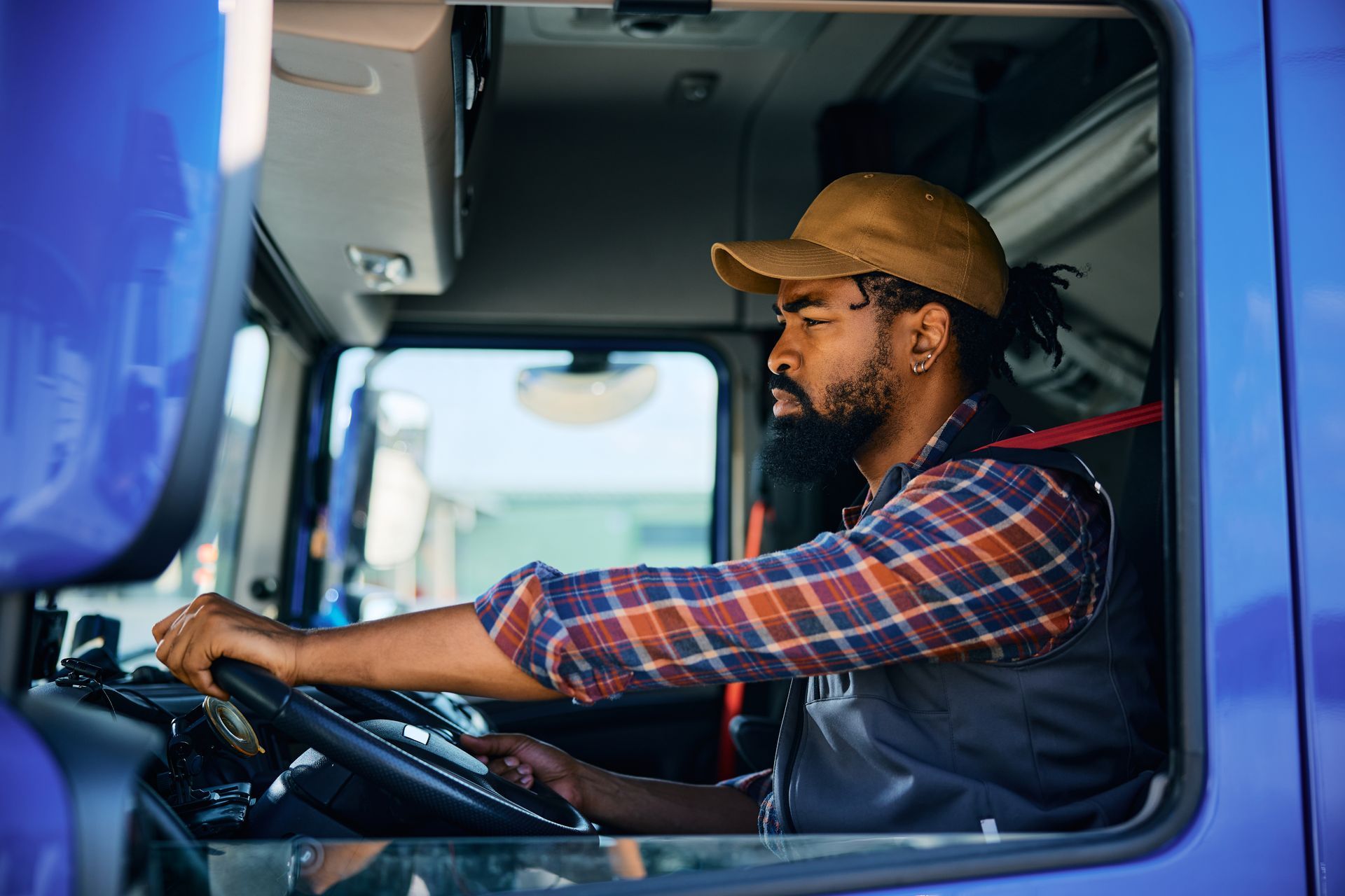 Truck driver in cap, driving a blue truck, looking forward with hand on the wheel.