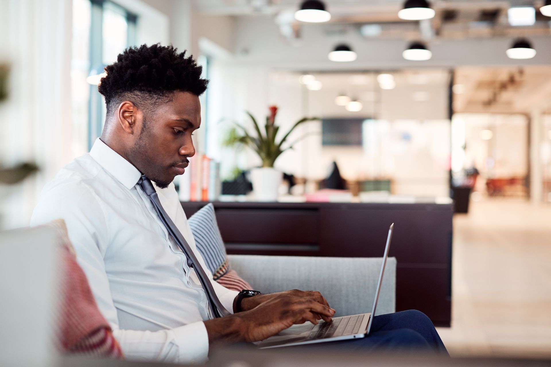 Man in white shirt and tie uses a laptop, seated on a couch in a modern office.