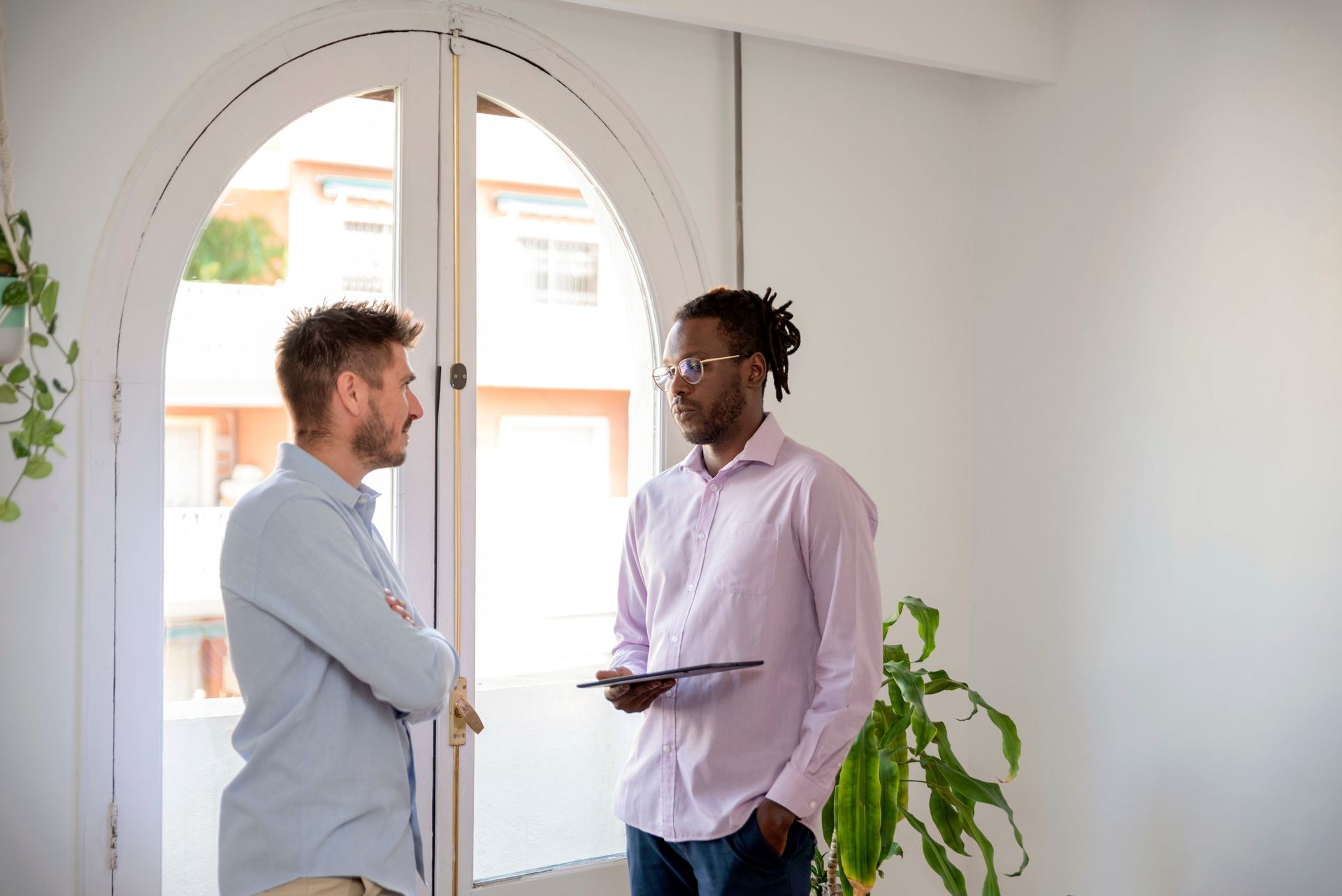Two men conversing near a window, one holding a tablet, in a bright room.