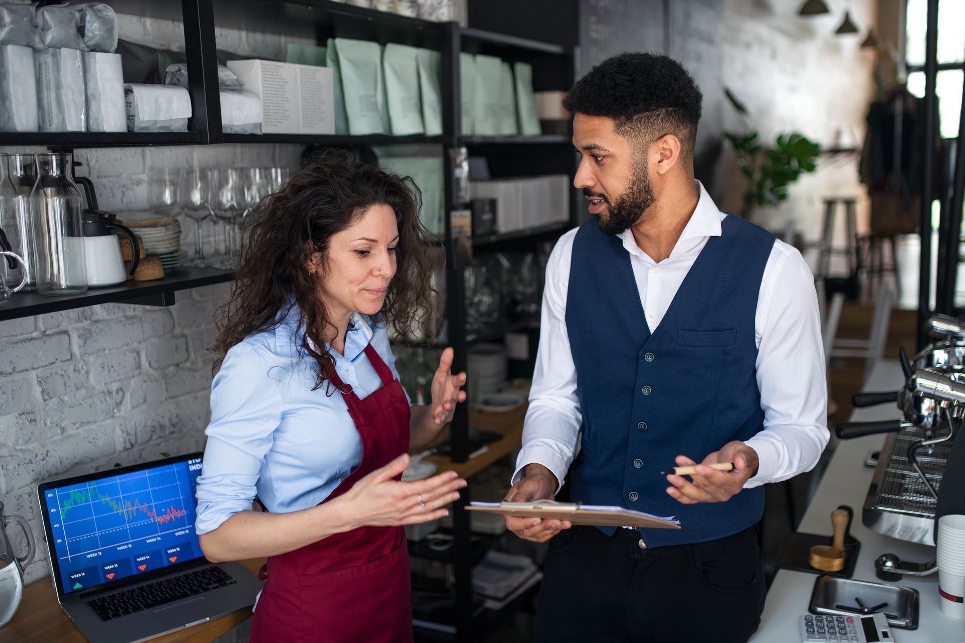 Barista and manager discussing business plans, holding a clipboard, in a cafe.