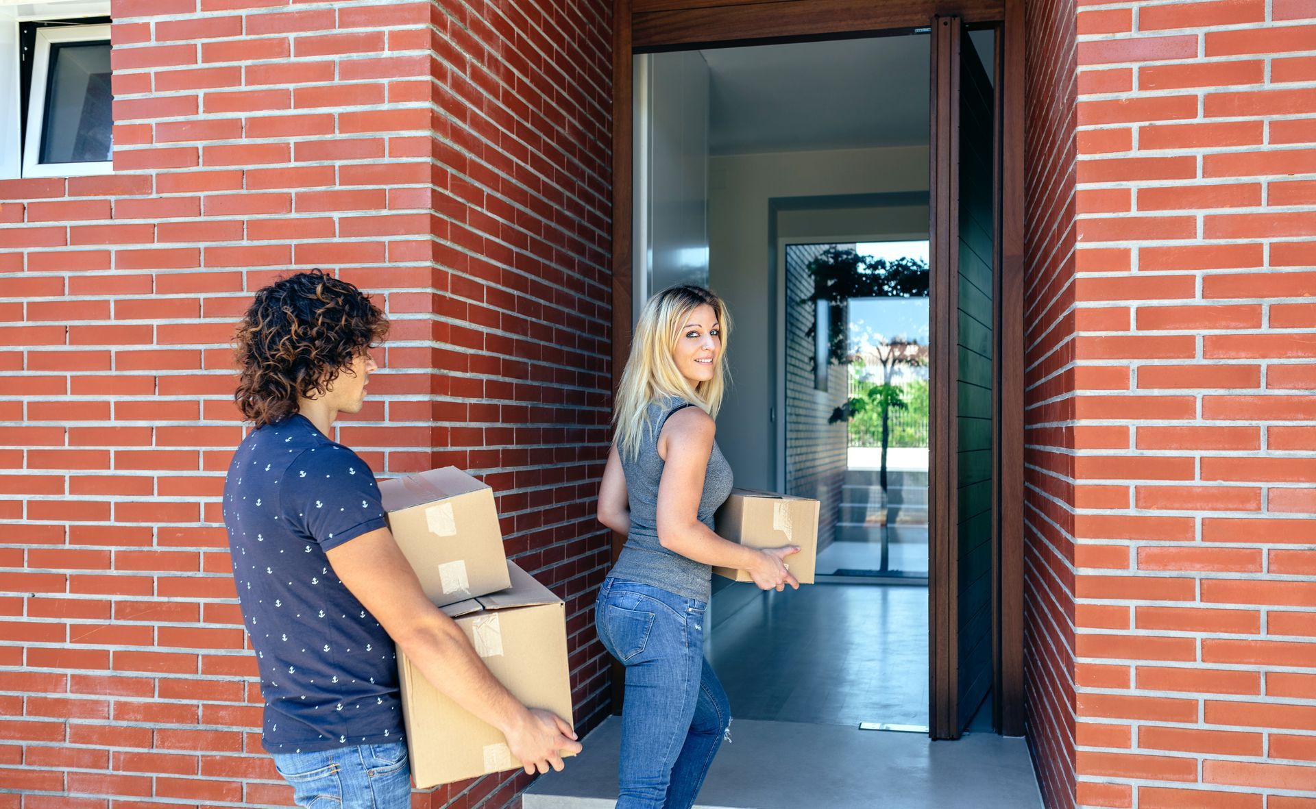 Couple carrying cardboard boxes into a brick home, entering through the open doorway.