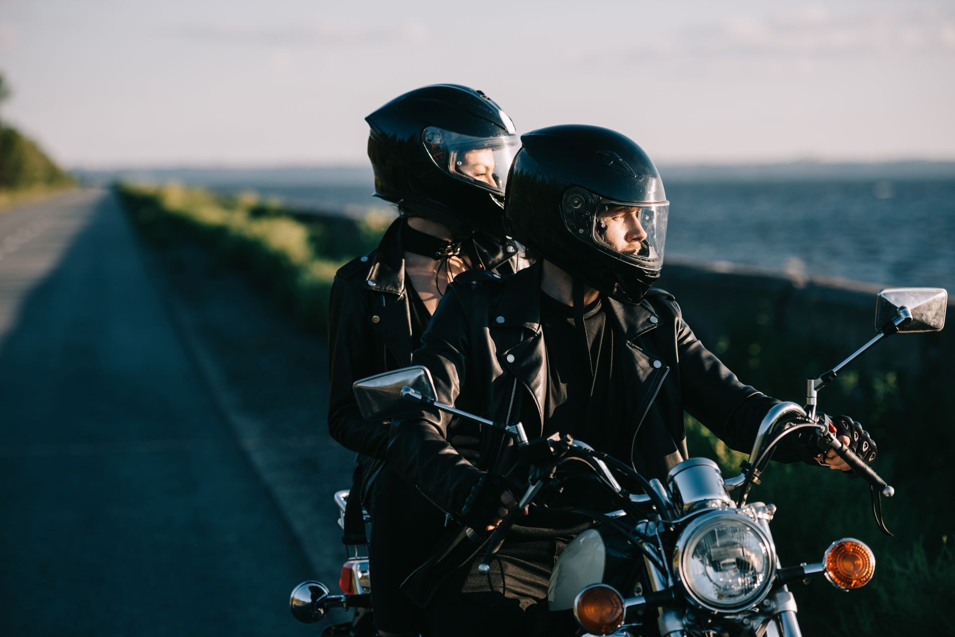 Two people on a motorcycle, wearing black helmets and jackets, riding down a road near a body of water.