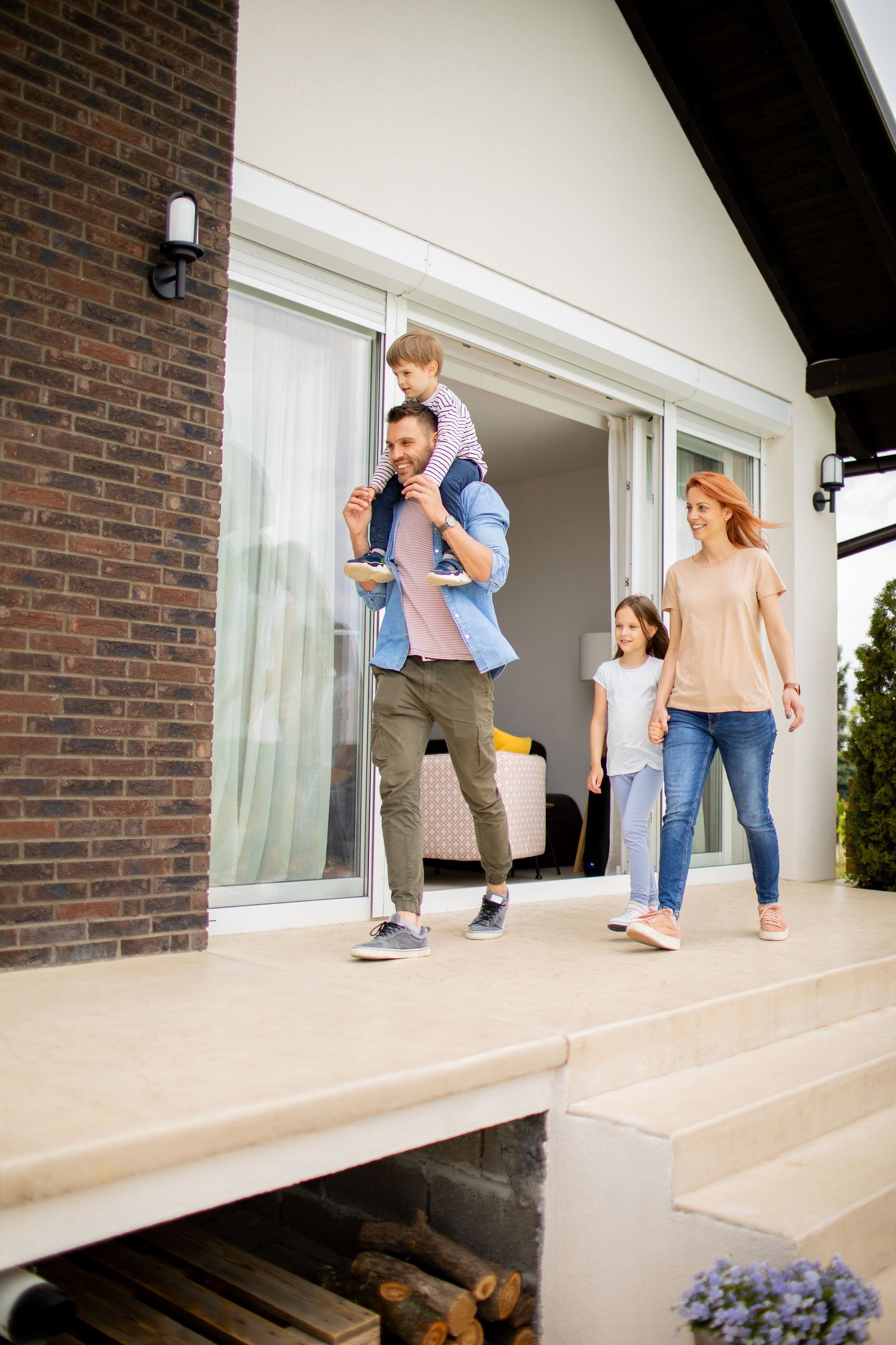 Family exiting a modern home. Father carries child on shoulders, mother and daughter walk beside them.