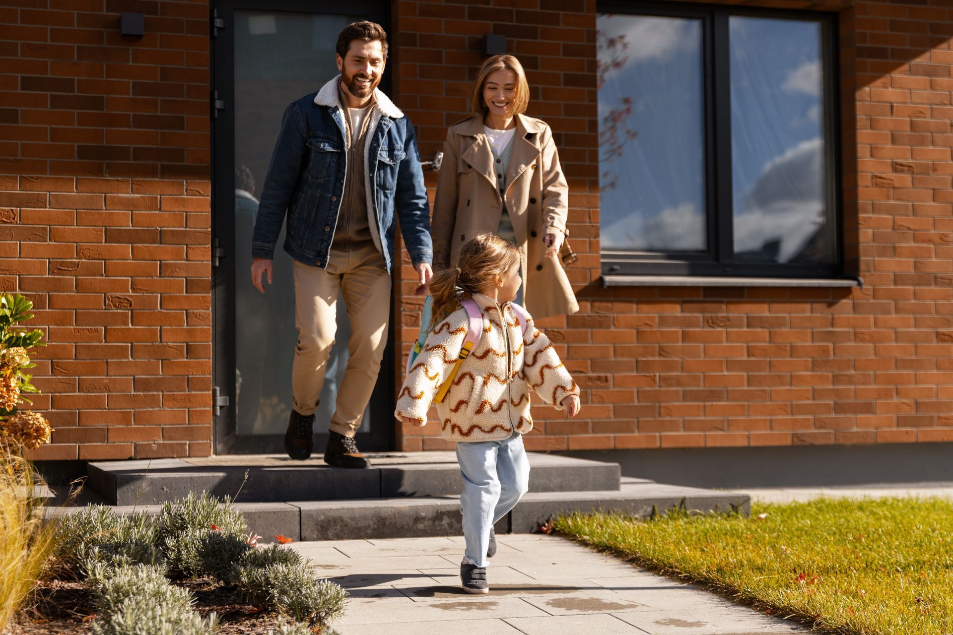 Family exiting a brick house. A child runs ahead while parents follow smiling.