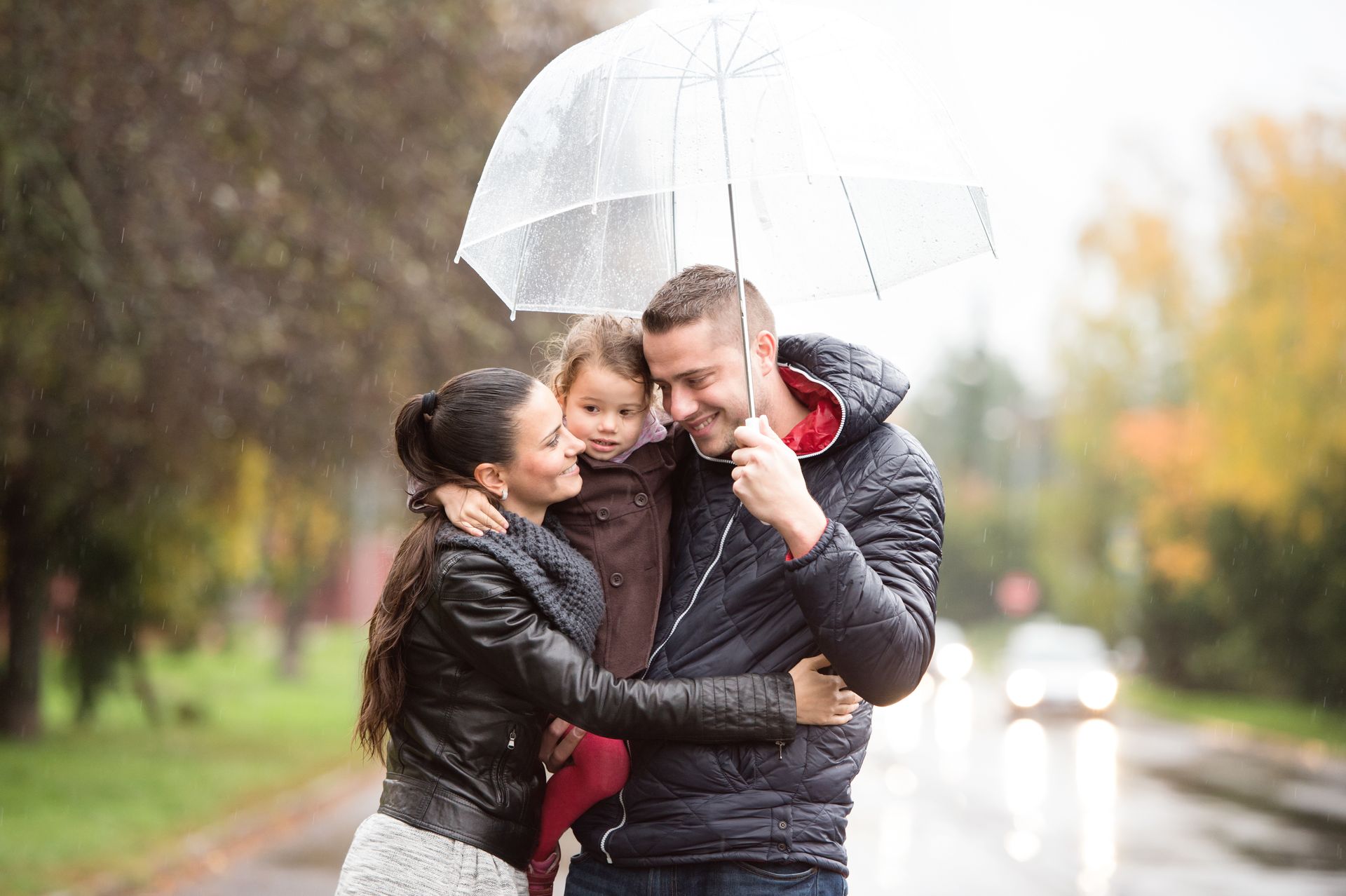 Family of three under a clear umbrella on a rainy day. Woman hugs a child; man smiles.