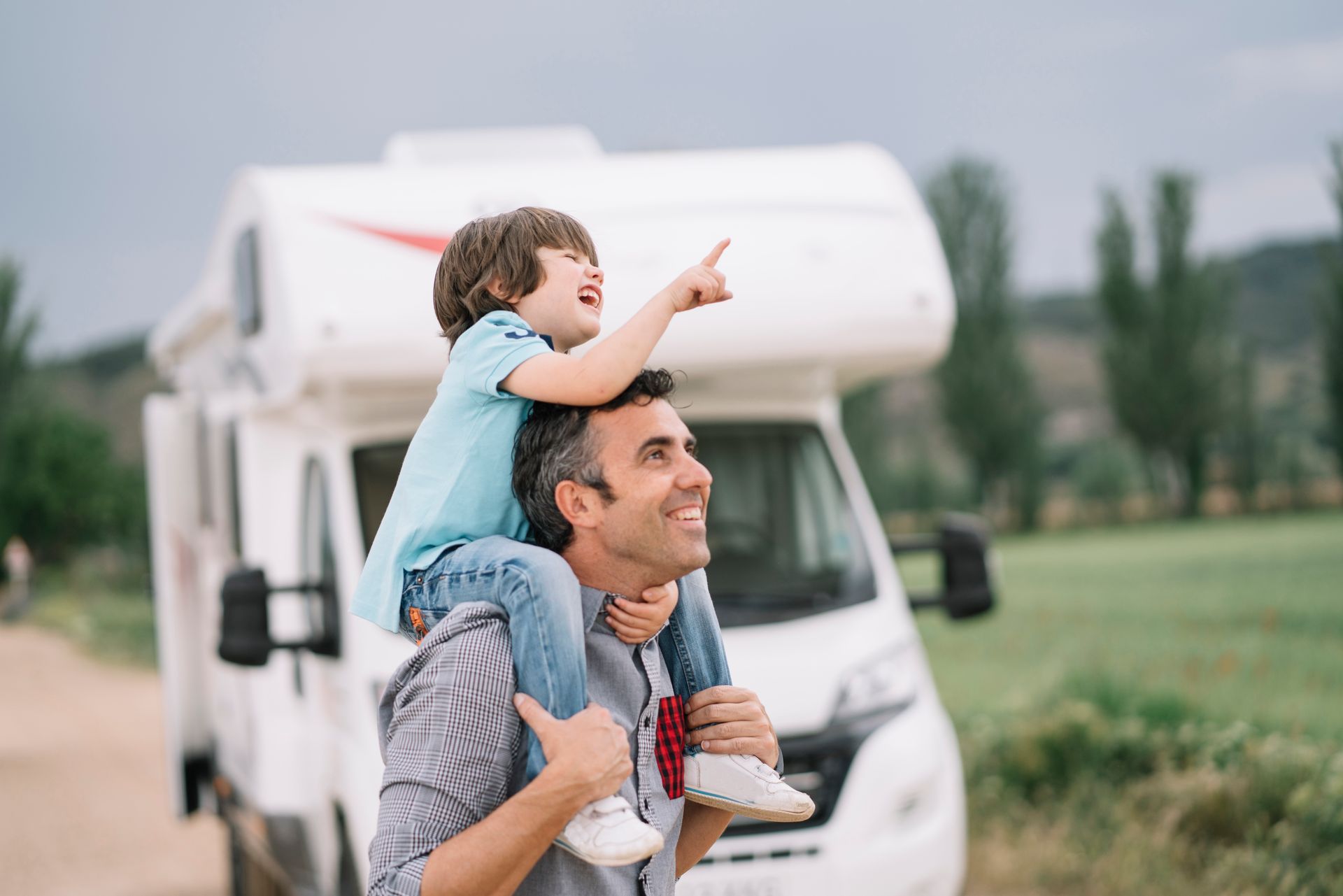 Man carrying a child on his shoulders, pointing towards the sky, with an RV in the background.