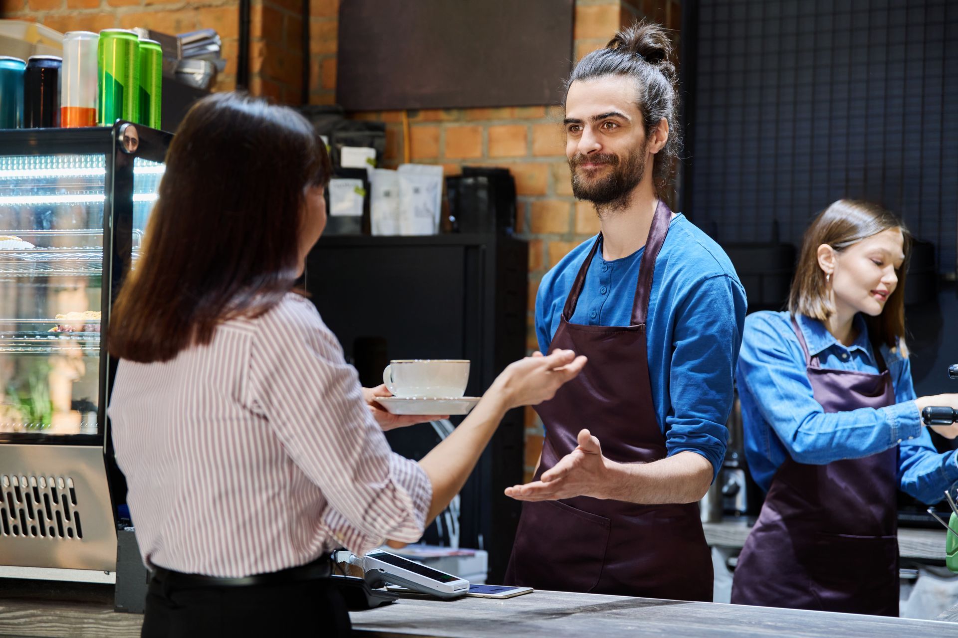 Customer receives order from barista at a coffee shop counter; second barista prepares something.