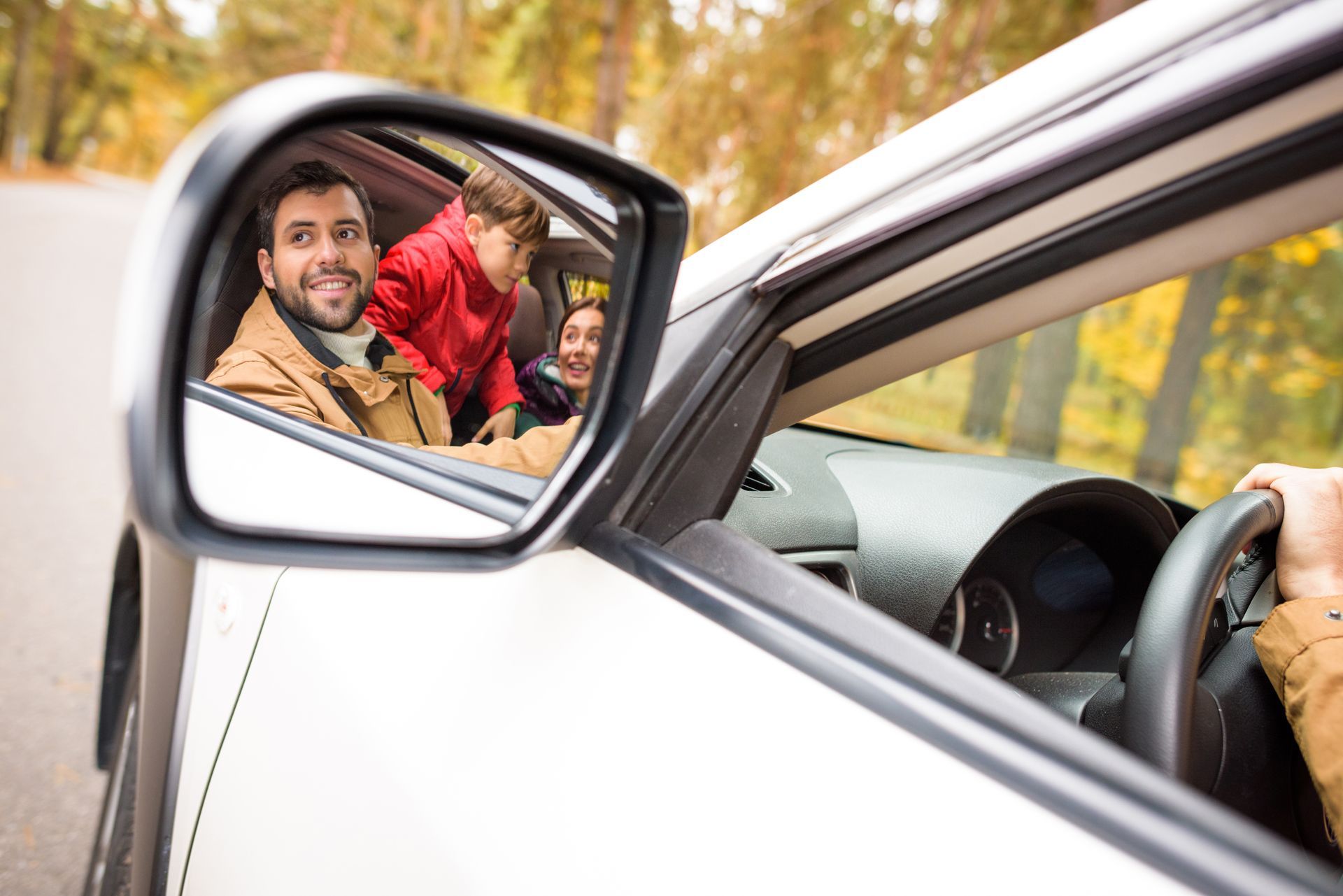 Side mirror reflects a man and two children in a car; driver's hand on steering wheel, outdoors with trees.