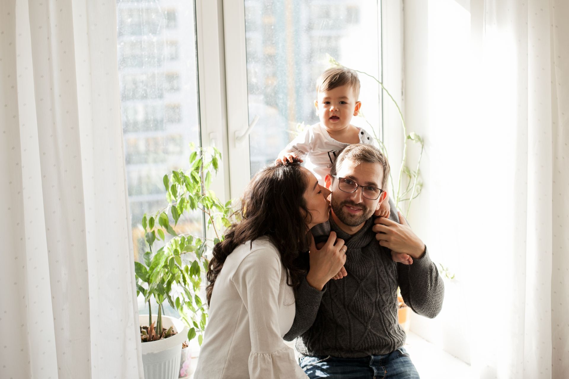 Family poses near a window. A person kisses another's cheek, while a child sits on the second person's shoulders.