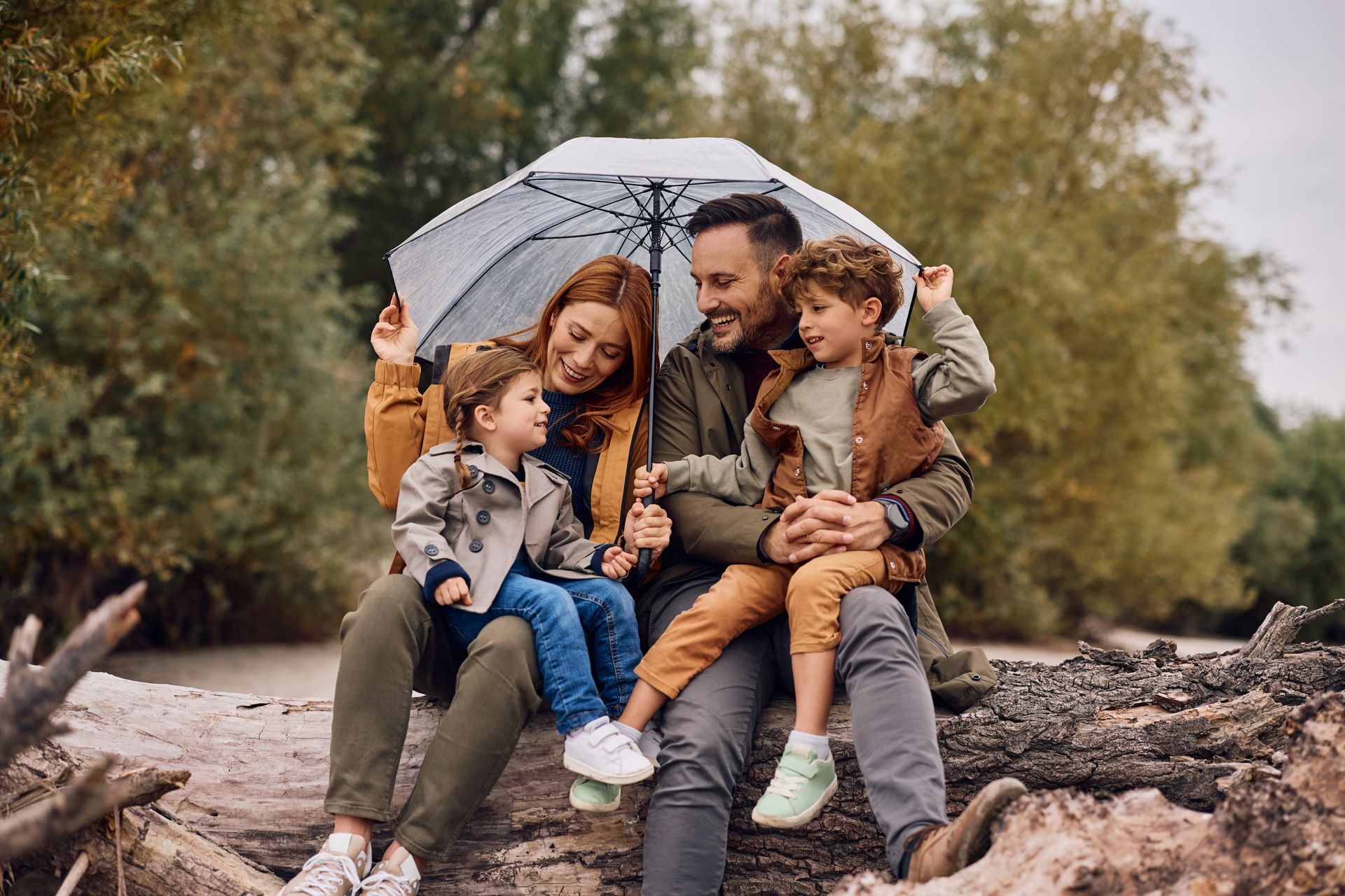 Family under umbrella, sitting on log, smiling outdoors. Trees in background, overcast day.
