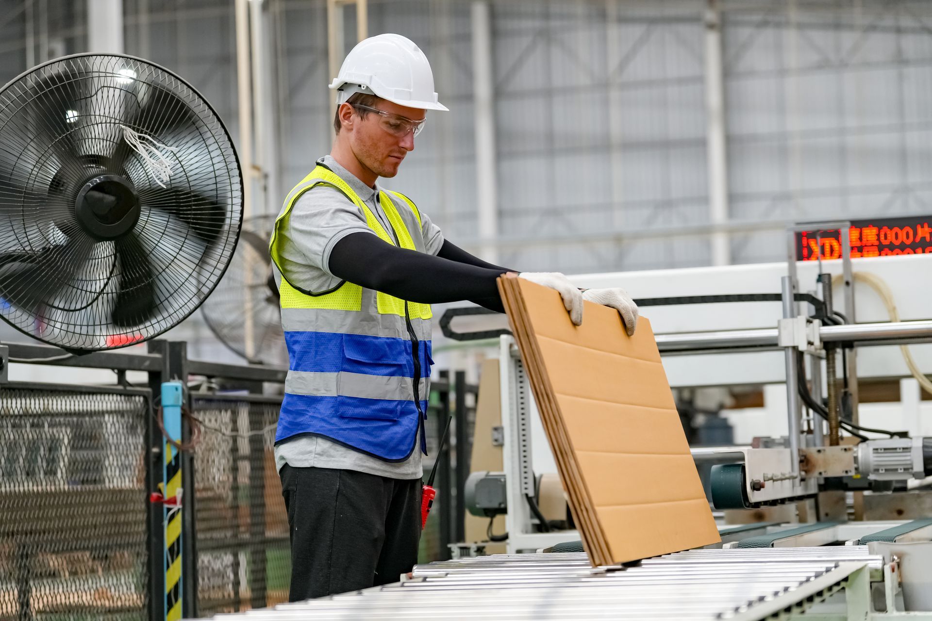 Factory worker in hard hat, safety vest, and gloves, loading cardboard onto a conveyor belt.