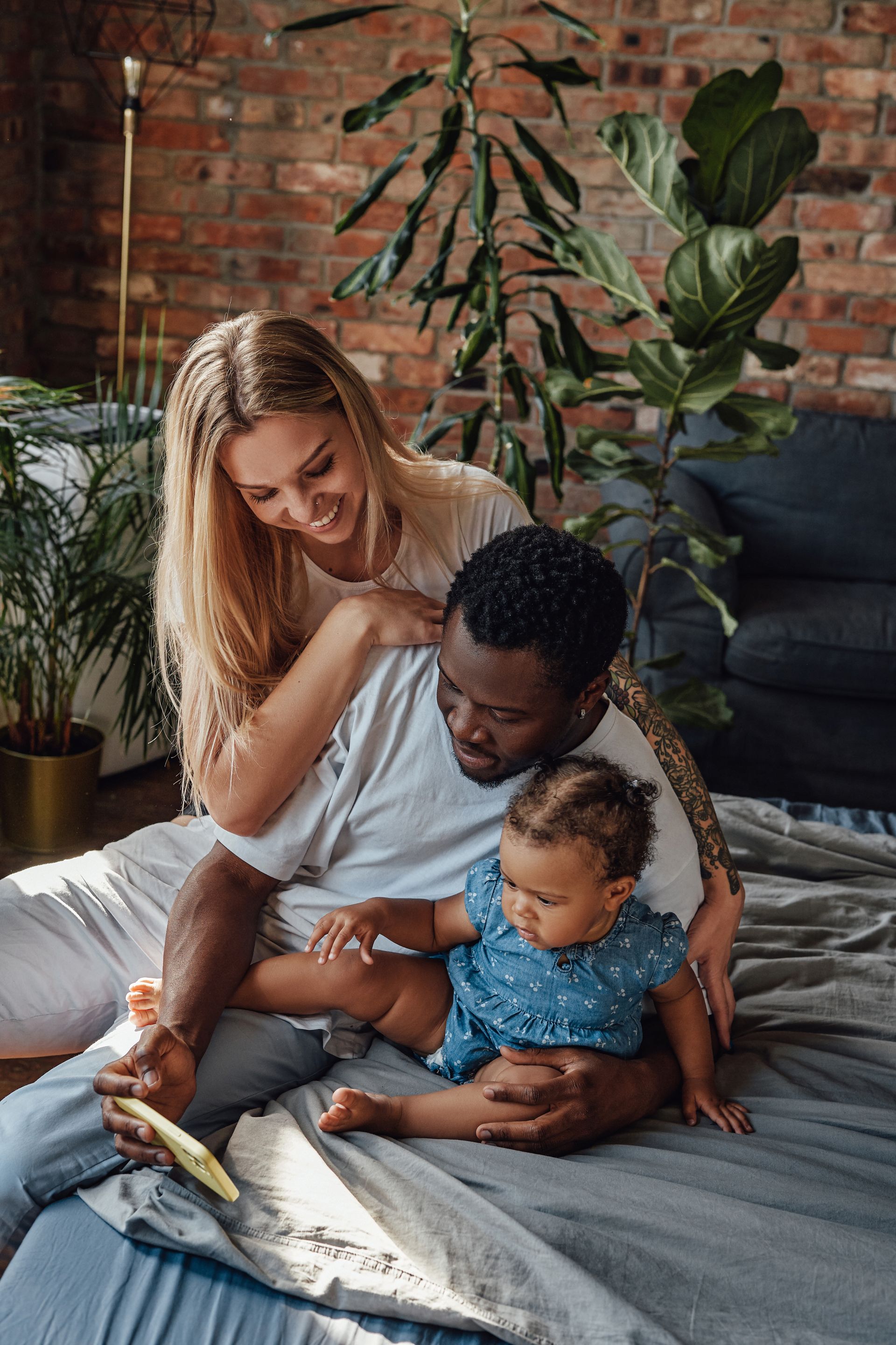 Family of three on a bed looking at a tablet, smiling. Brick wall, plants in the background.