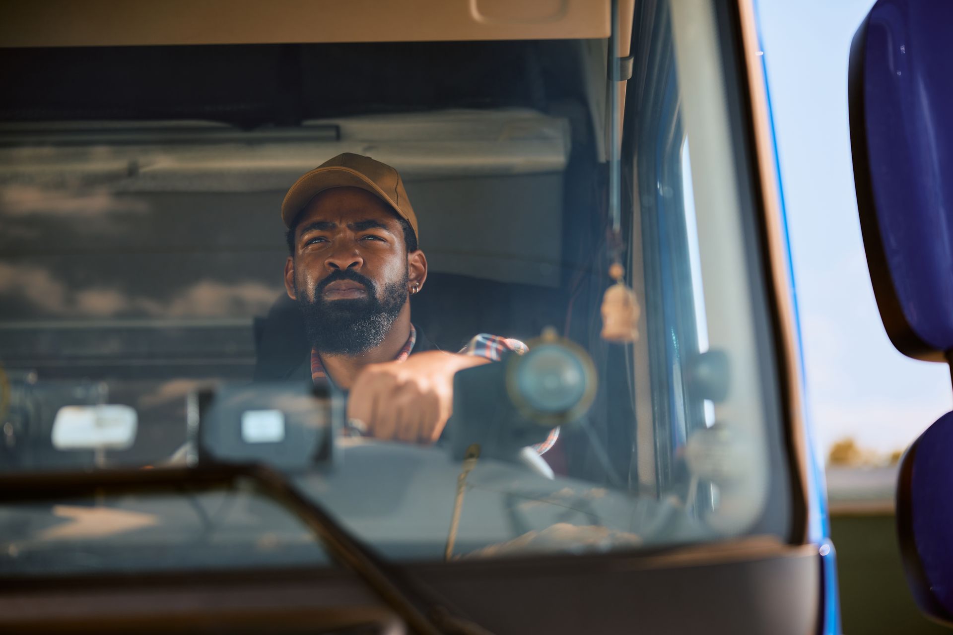Truck driver looking ahead through the windshield, wearing a baseball cap and dark shirt.