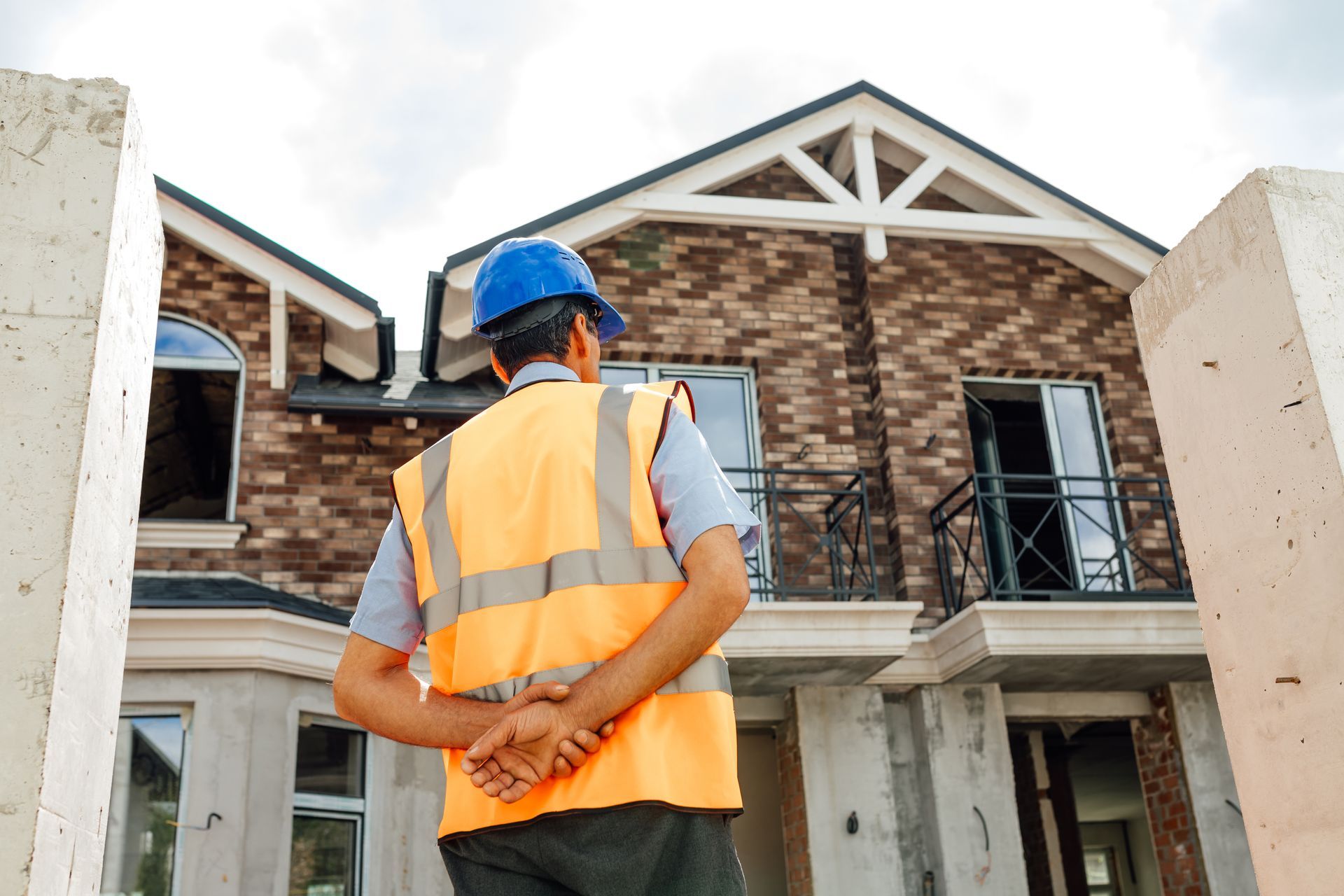 Construction worker in safety vest and hard hat surveying a partially built brick house.