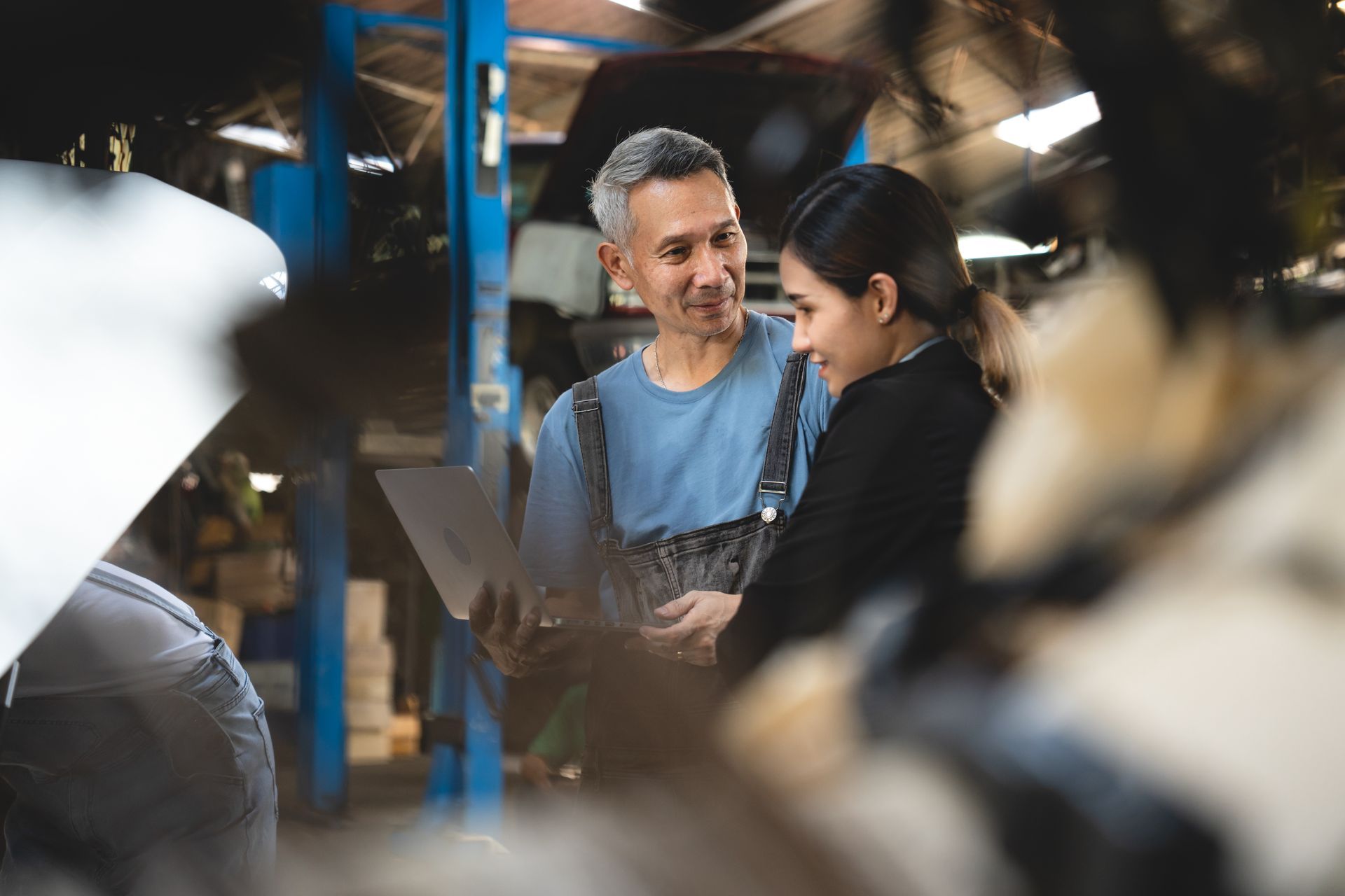 Mechanic and woman looking at a laptop in a garage. Man in overalls, woman in black jacket.