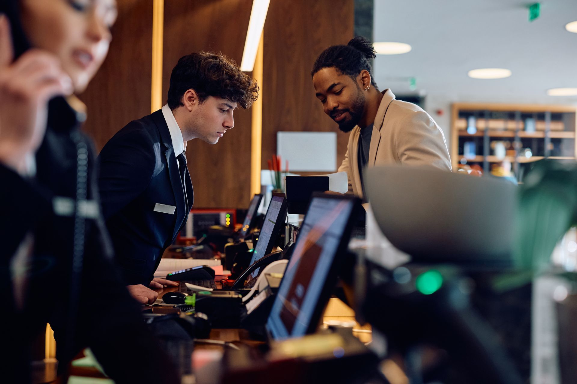 Hotel check-in: Desk clerk helps a guest, another person answers a phone. Warm, wooden interior.