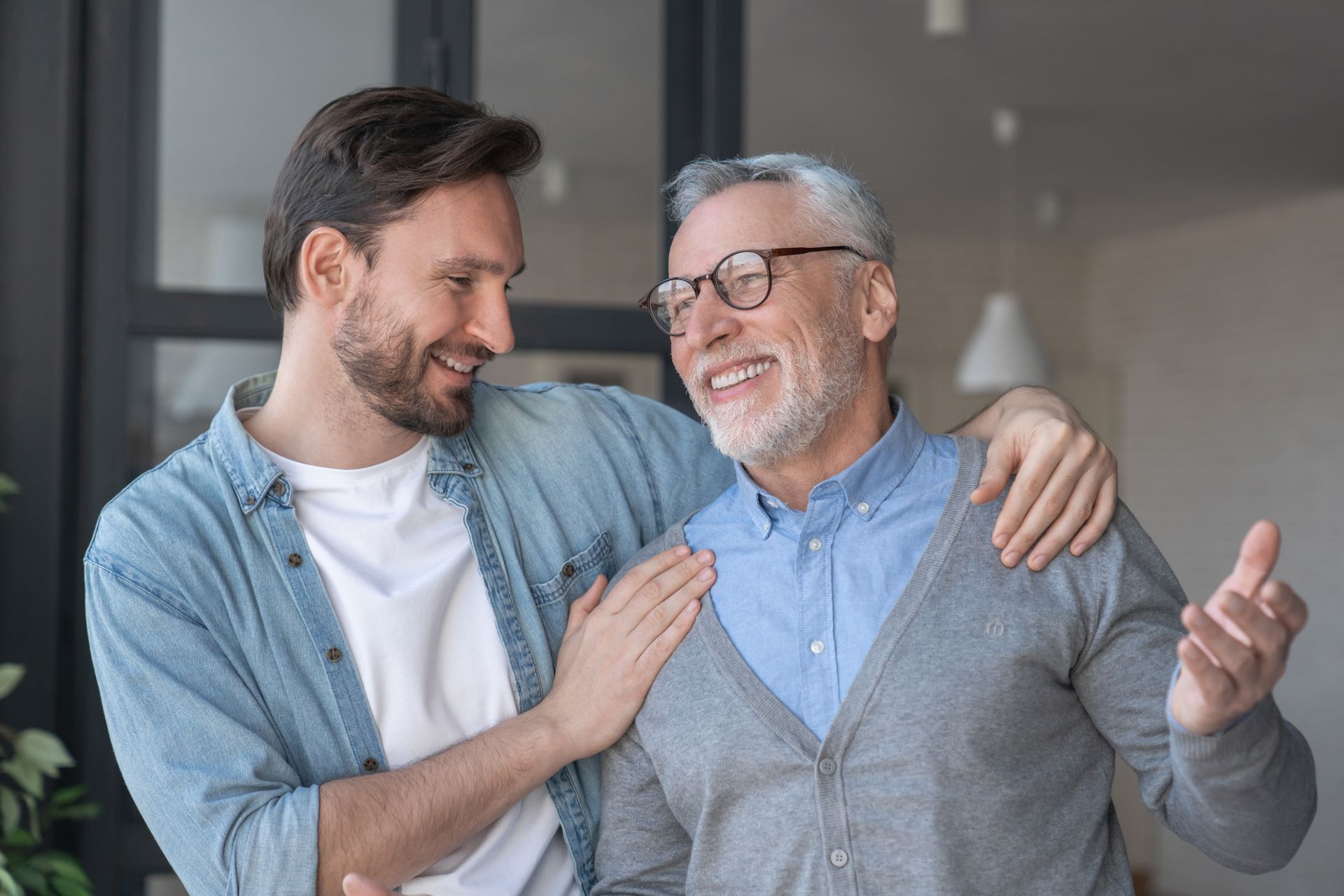 Man with beard embraces older man wearing glasses, both smiling. Indoors.