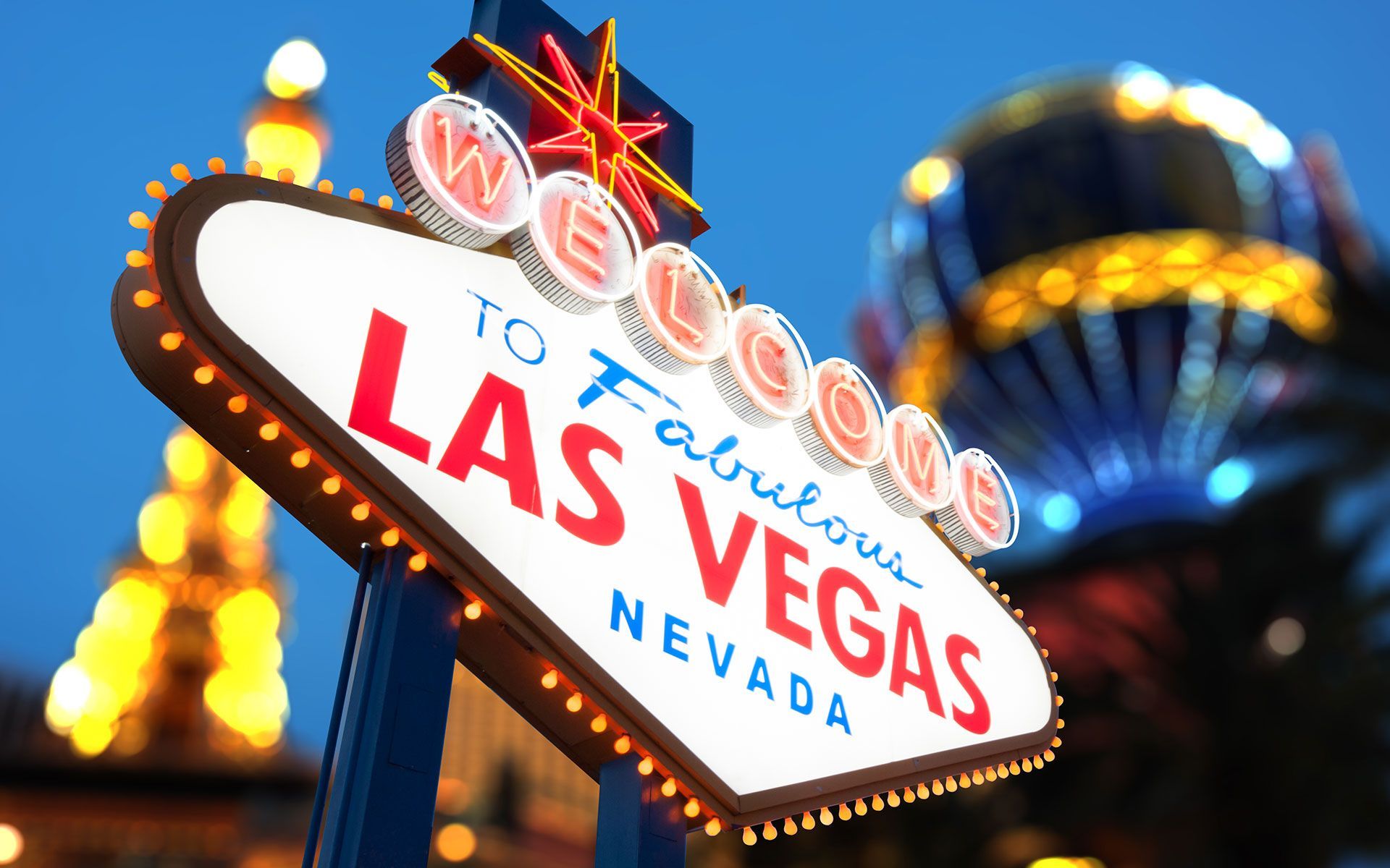 Las Vegas welcome sign against a blue sky with blurred lights in the background.