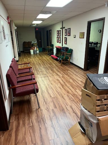 A long hallway with wooden floors , chairs , boxes and a cross on the wall.