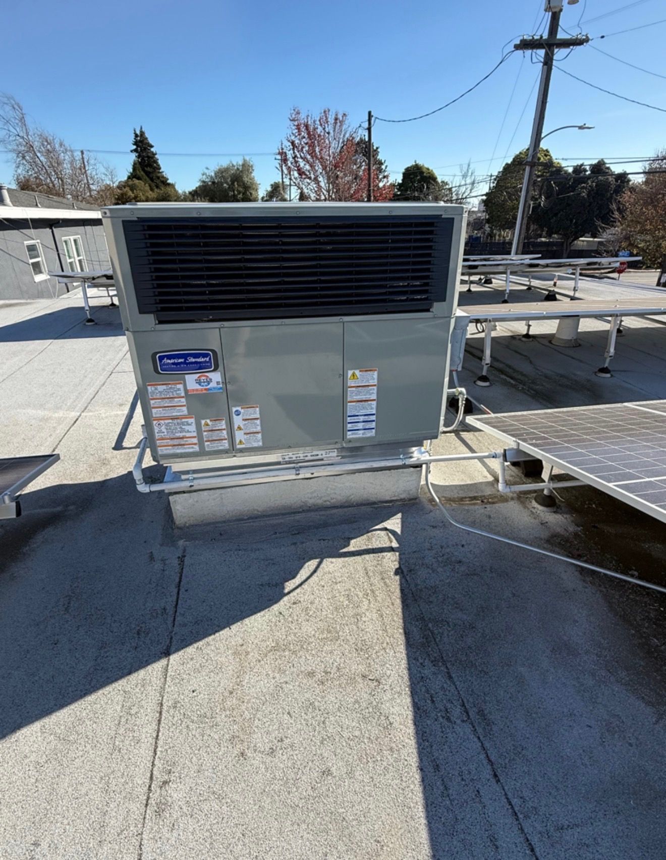 A gray HVAC unit sits on a flat rooftop on a sunny day, with solar panel racks visible to the right.