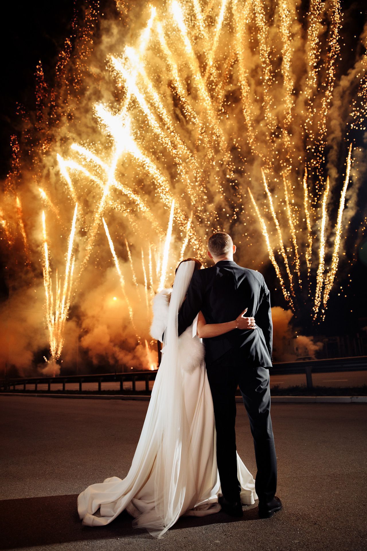 A bride and groom are standing in front of a fireworks display.