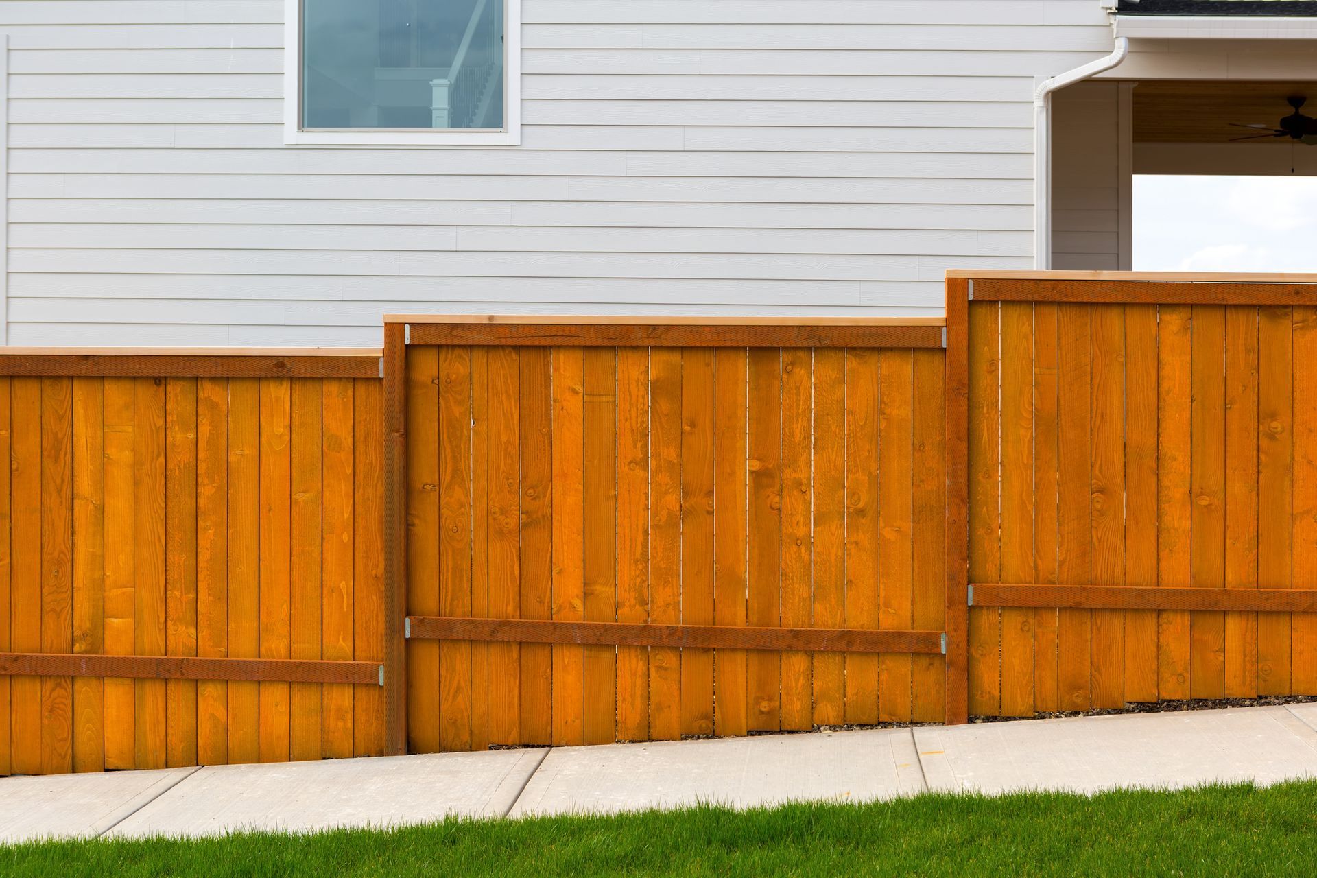 A stepped, stained wooden privacy fence stands in front of a white-sided house, built along a concrete sidewalk.