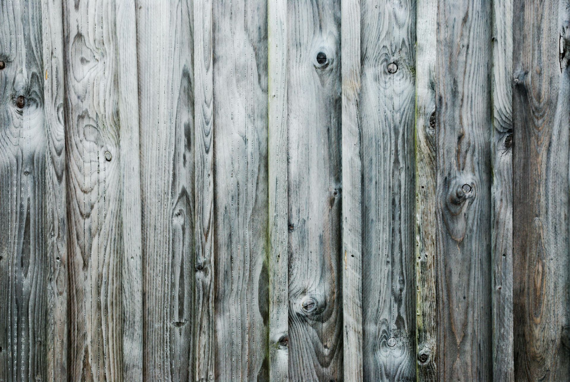 A vertical arrangement of weathered, grey wooden planks with visible grain, knots, and a rustic texture.