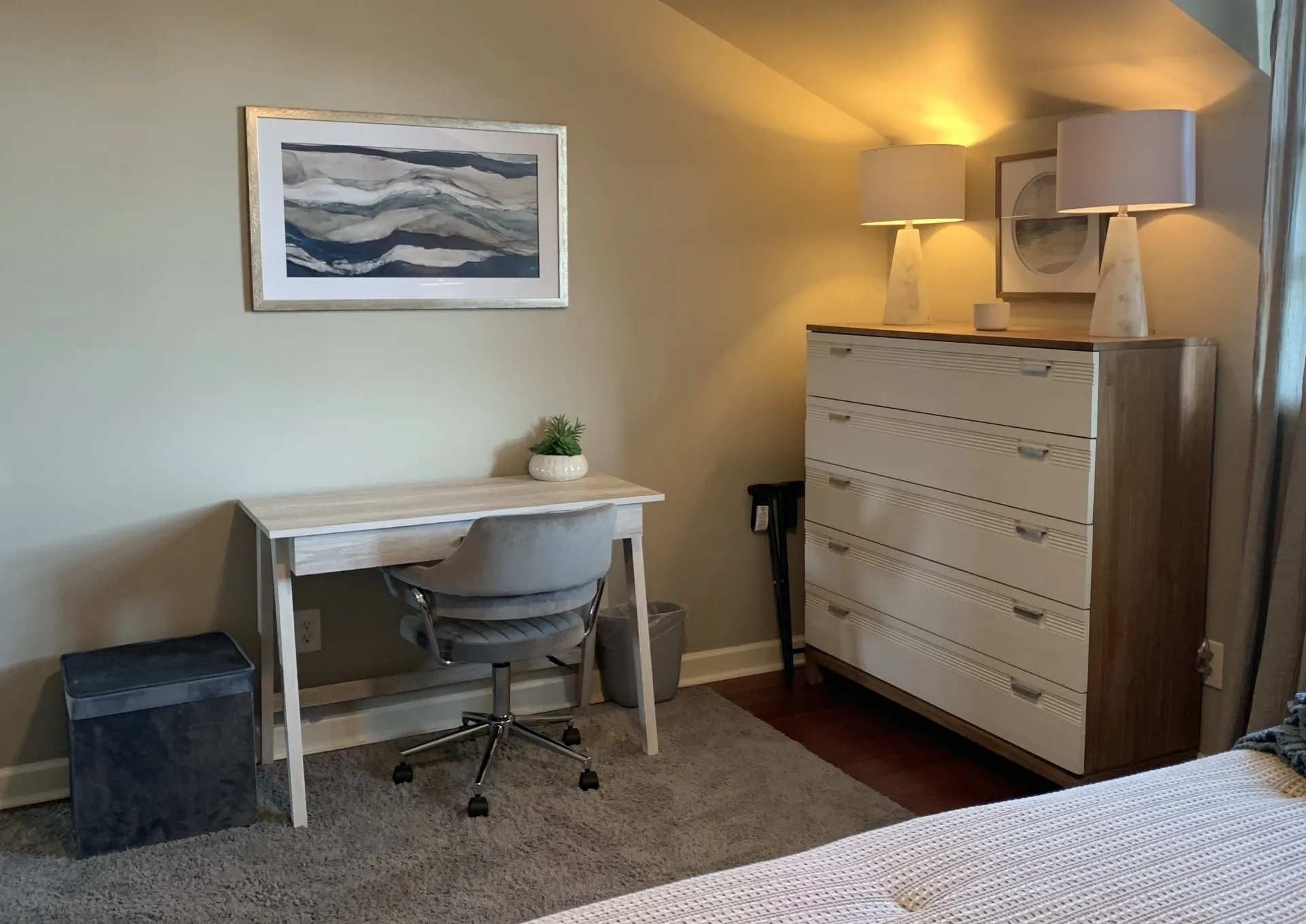 A bedroom with a desk , chair , dresser and lamps.