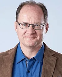 A man wearing glasses and a blue shirt is smiling for the camera.