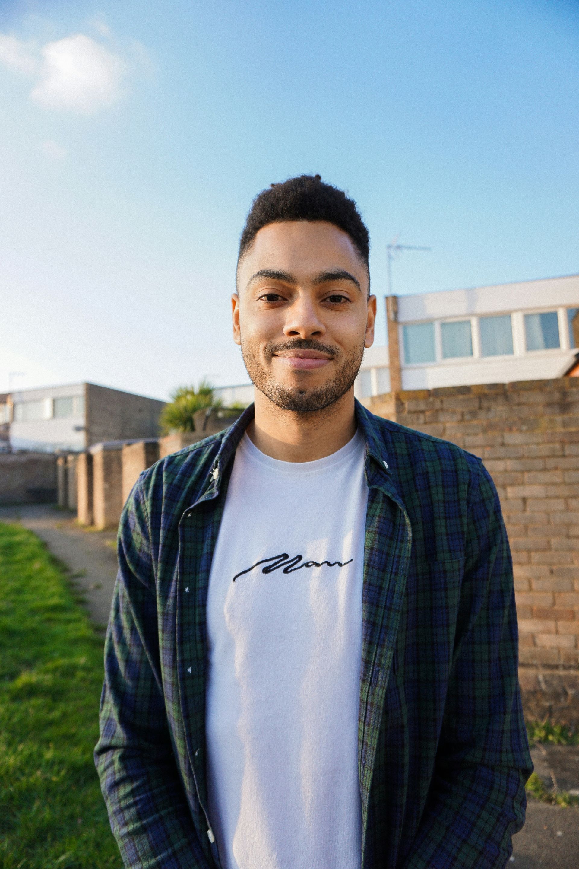 A man wearing a plaid shirt and a white t-shirt is standing in front of a brick wall.