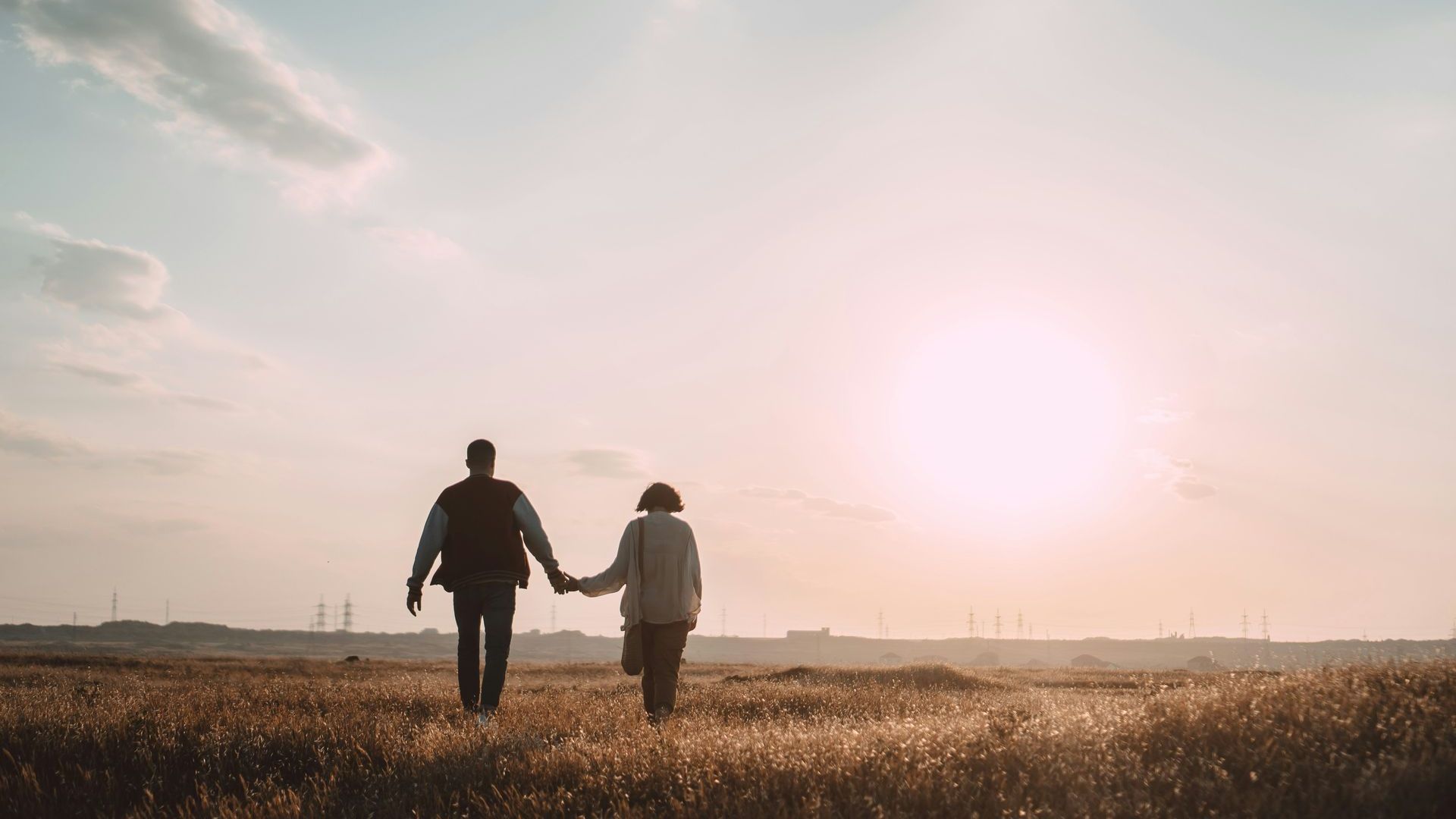A man and a woman are holding hands while walking through a field at sunset.