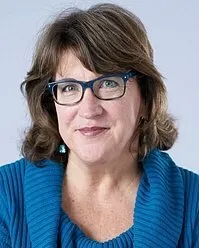 A woman wearing glasses and a blue sweater is smiling for the camera.