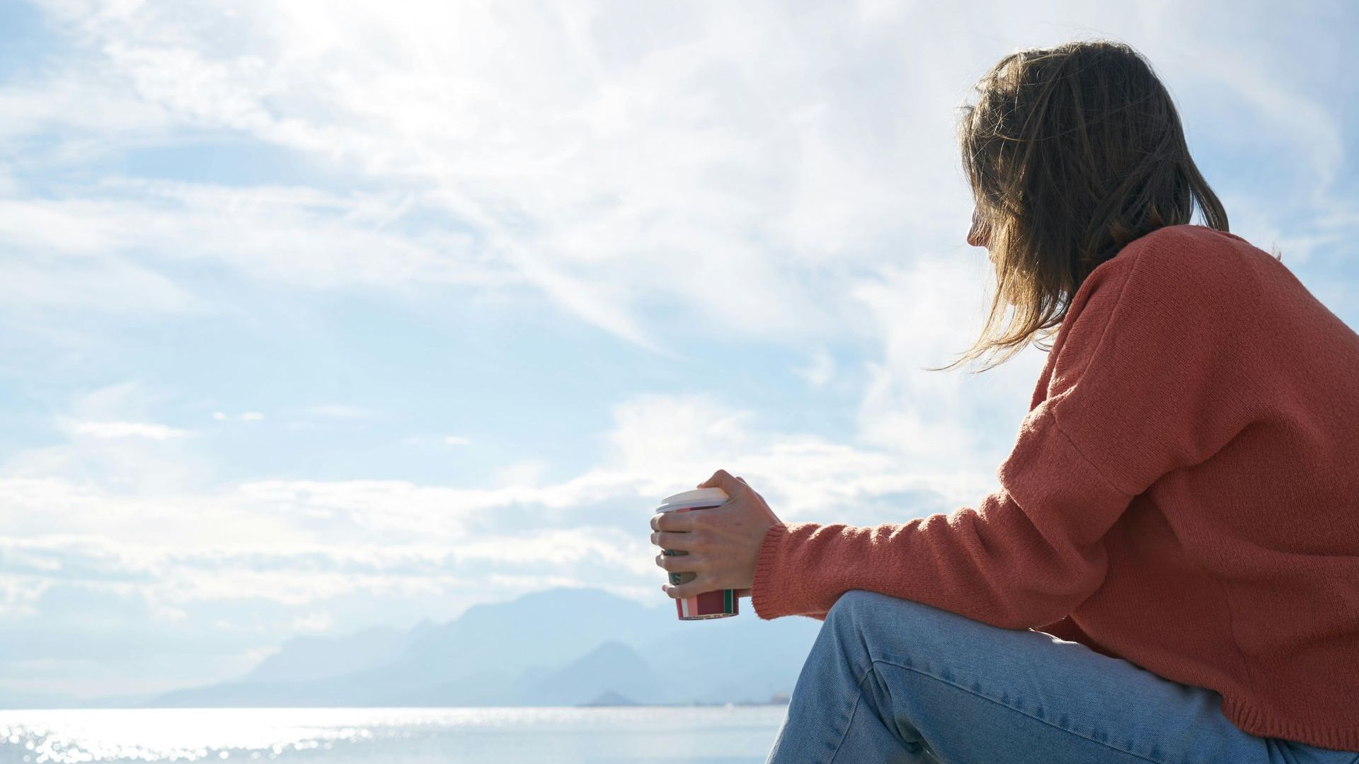 A woman is sitting on the edge of a cliff holding a cup of coffee.