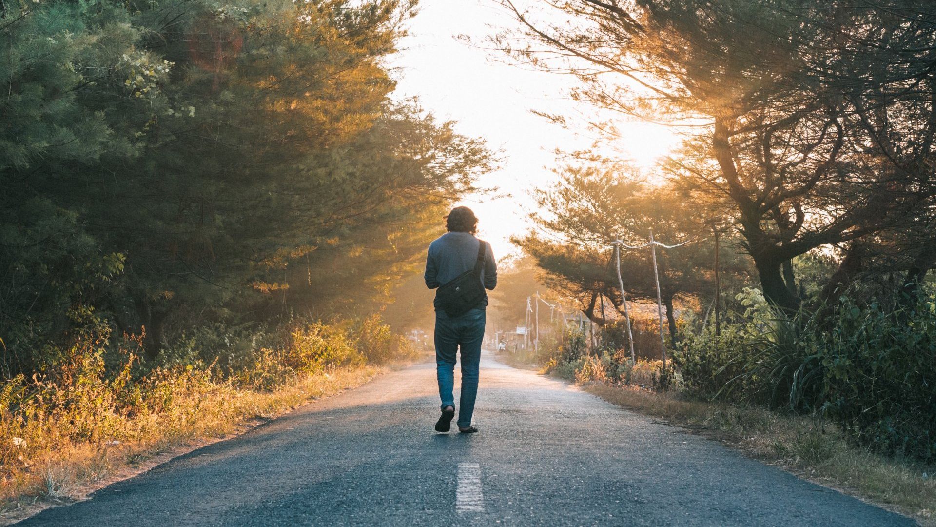 A man is walking down a road surrounded by trees at sunset.