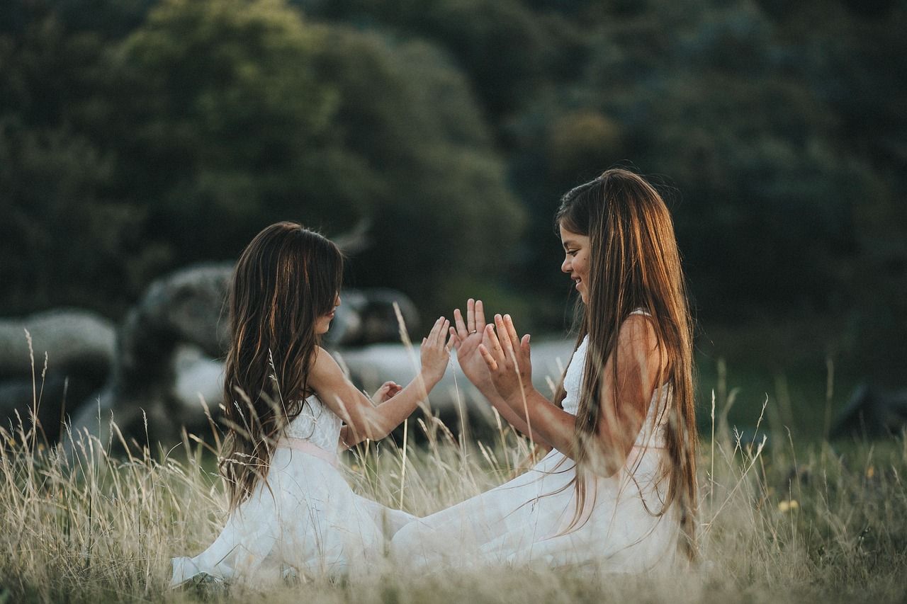 Two little girls are sitting in the grass playing with their hands.