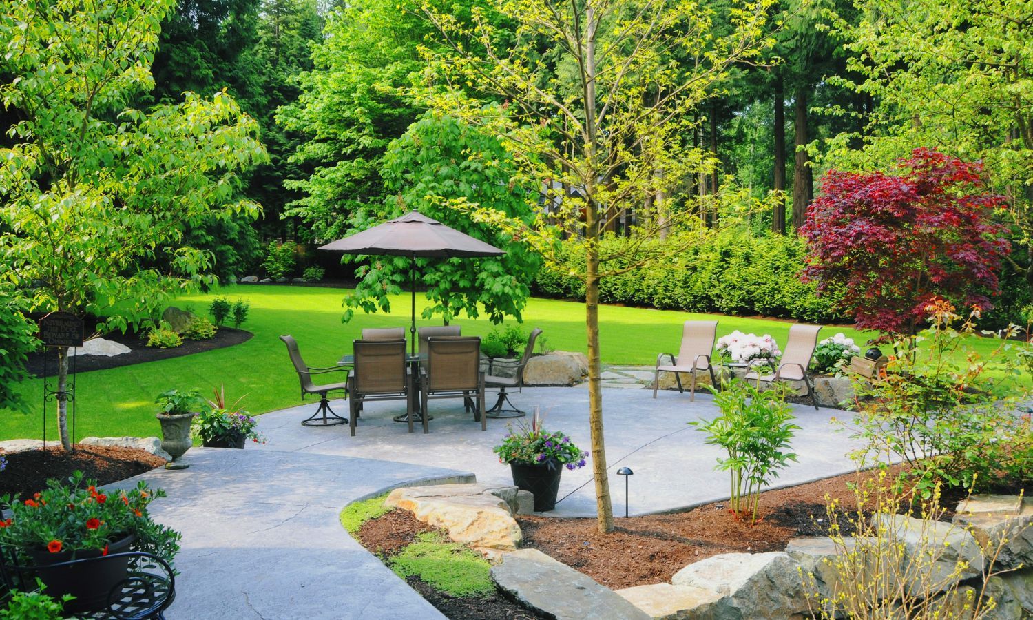 A lush garden patio with furniture, an umbrella, surrounded by trees and greenery.