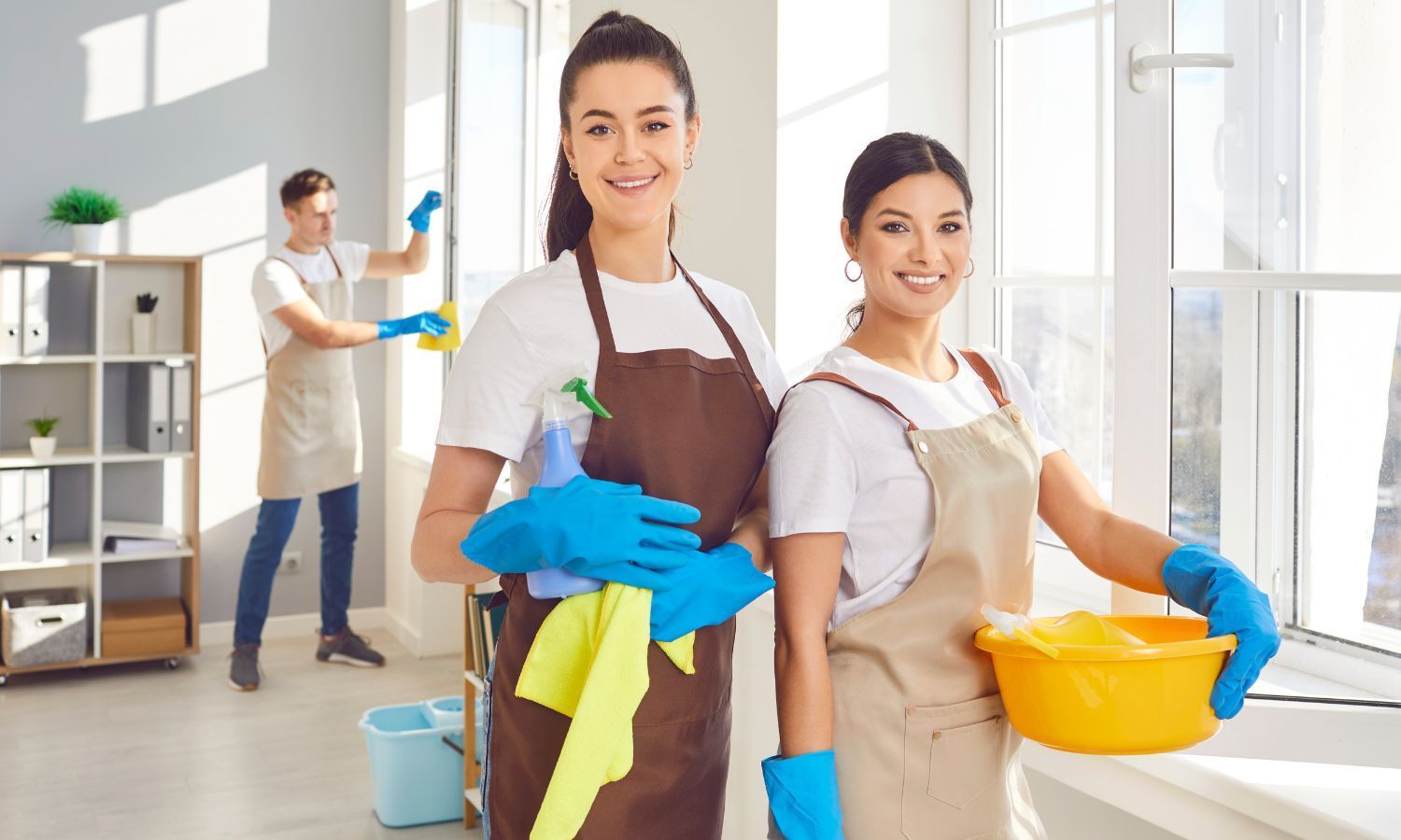 Three cleaning staff smiling, two in foreground holding cleaning supplies, one cleaning windows in background.