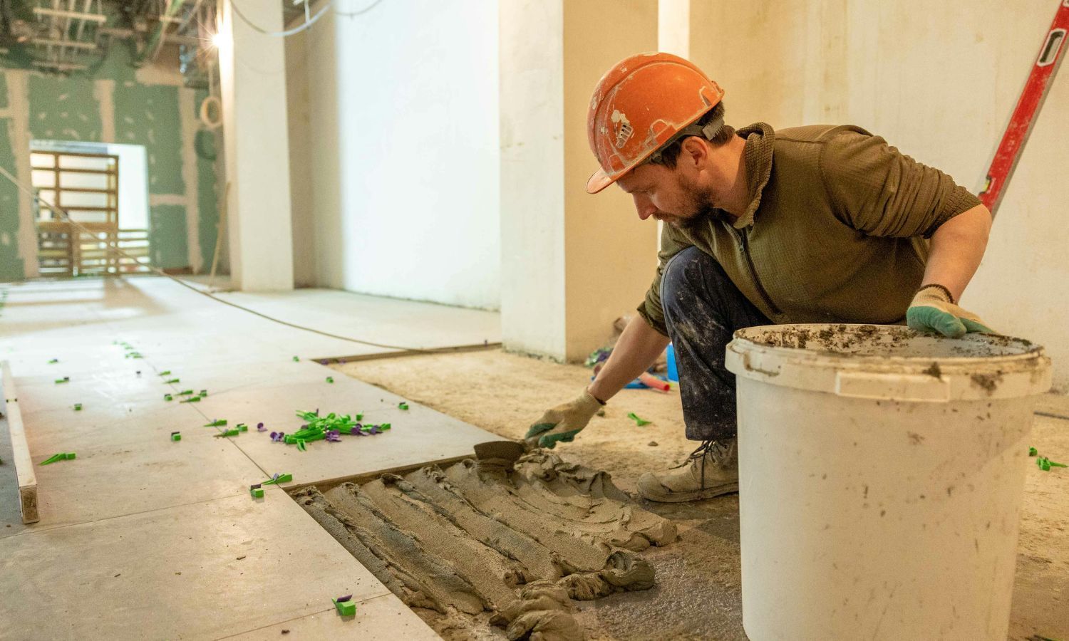 Construction worker in a hard hat spreading mortar on a floor, installing tiles in a room.