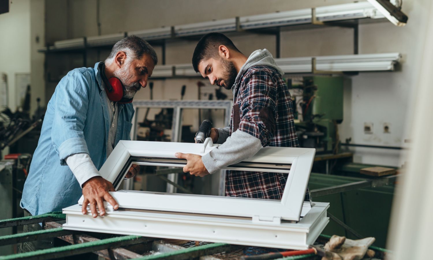 Two men assembling a window frame in a workshop, one older with ear protection, the other younger, both focused.