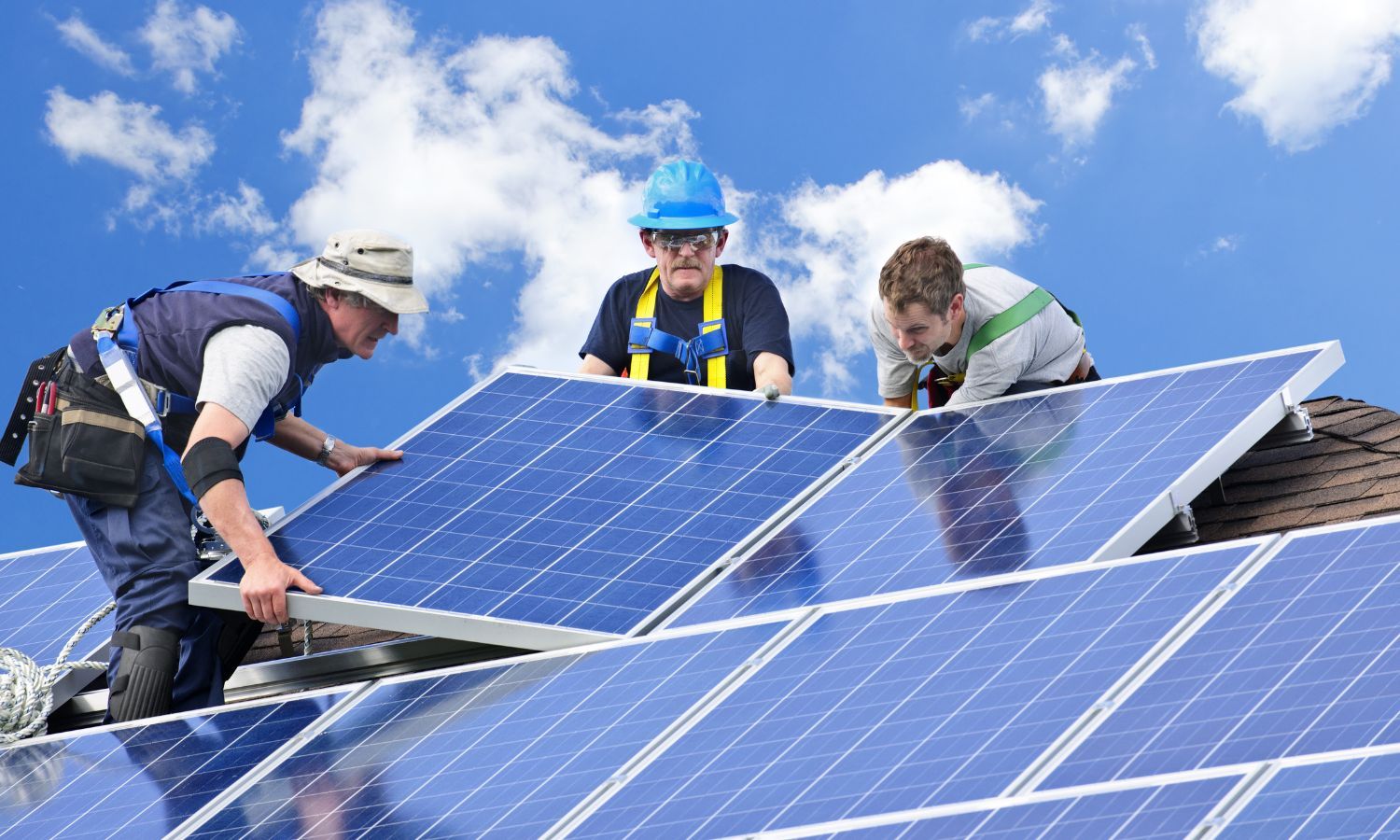 Three workers install solar panels on a rooftop against a blue sky.