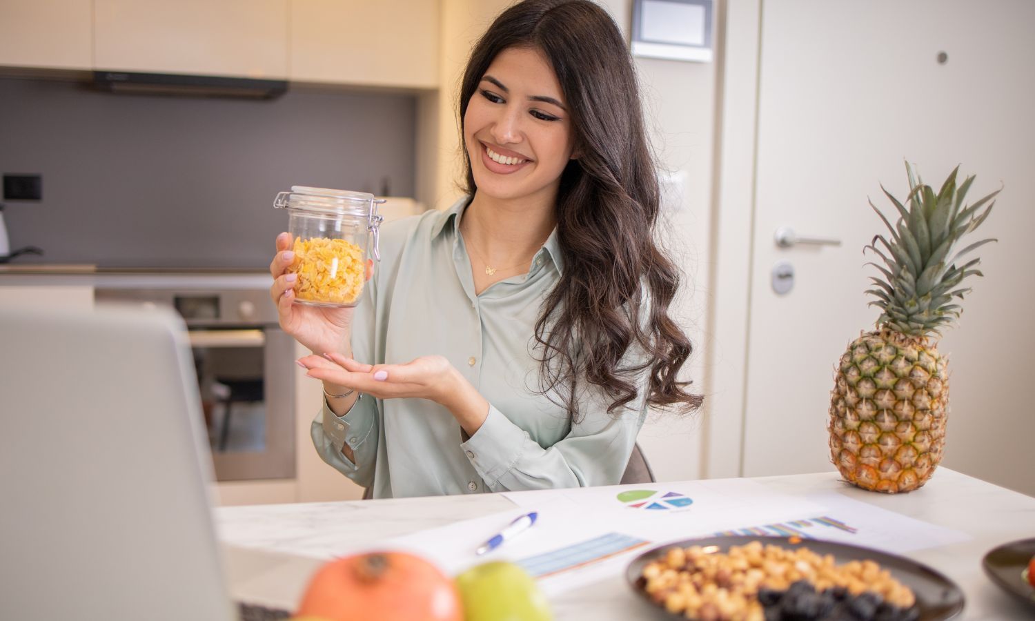 Woman holding jar of food, smiling, with fruit and laptop in a kitchen.