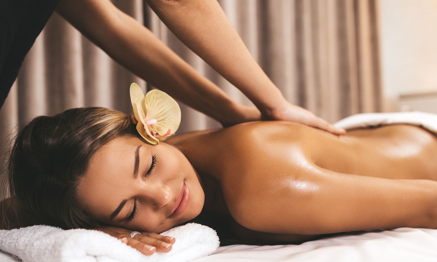 Woman receiving a back massage in a spa, with flower in hair, eyes closed, hands on back.