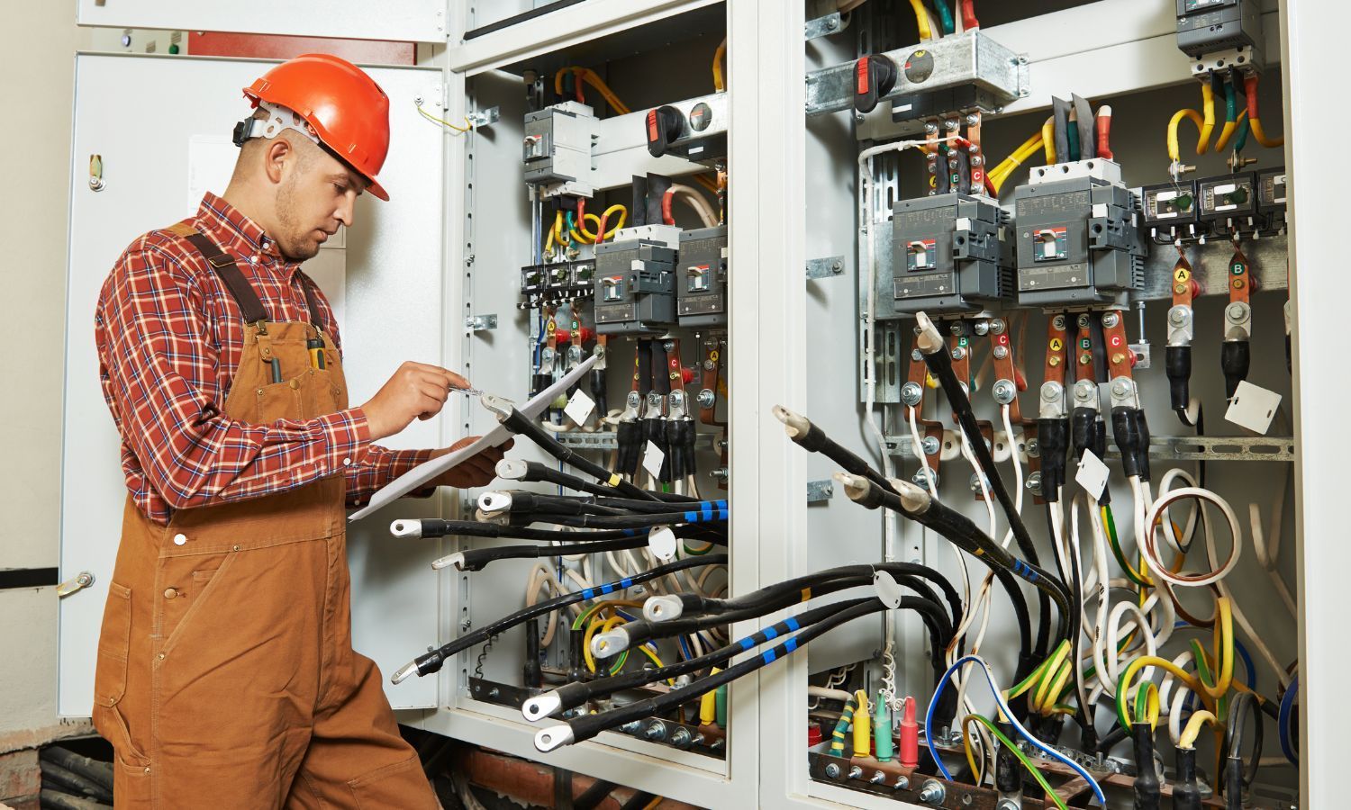 Electrician in orange hard hat and overalls inspects wiring inside an electrical panel.