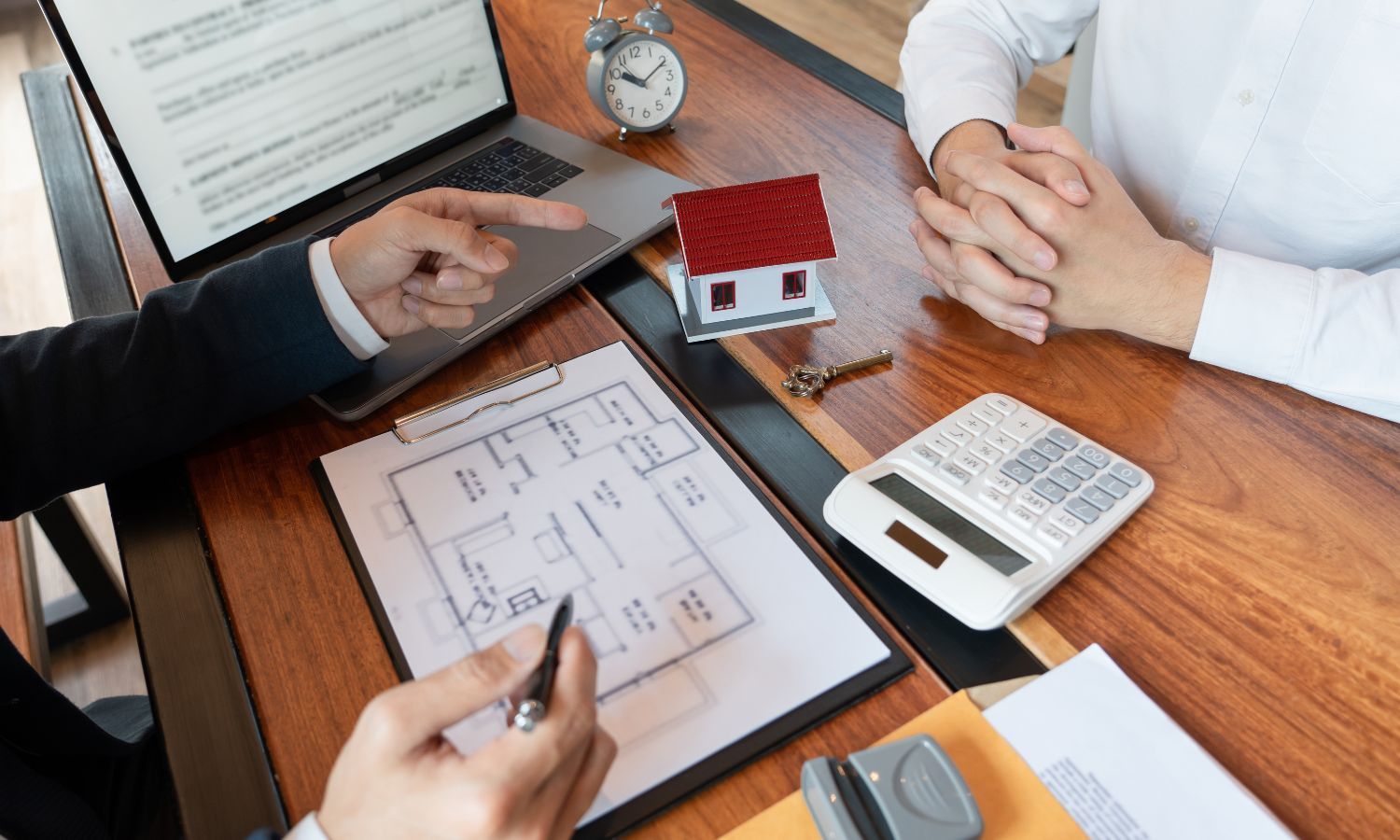Real estate agent pointing at floor plan, discussing with a client, model house and calculator on desk.