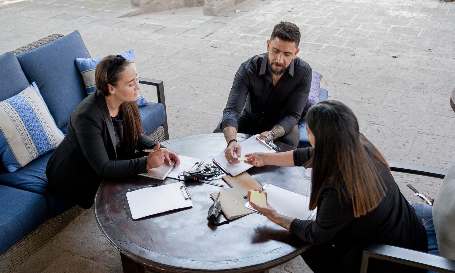 Three people at a round table outside, looking at papers. Two women and a man, possibly discussing designs.
