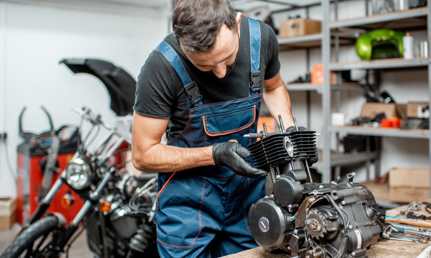 Mechanic in overalls working on a motorcycle engine in a workshop, wearing black gloves.