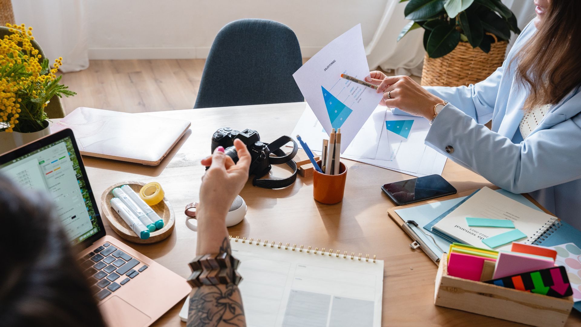 Two women reviewing charts at a desk, one points with pen. Laptop, camera, and stationery present.